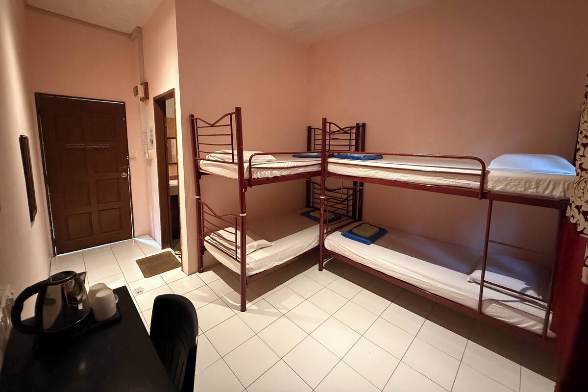 A dormitory room with bunk beds, a door, a table with a kettle, and a chair.