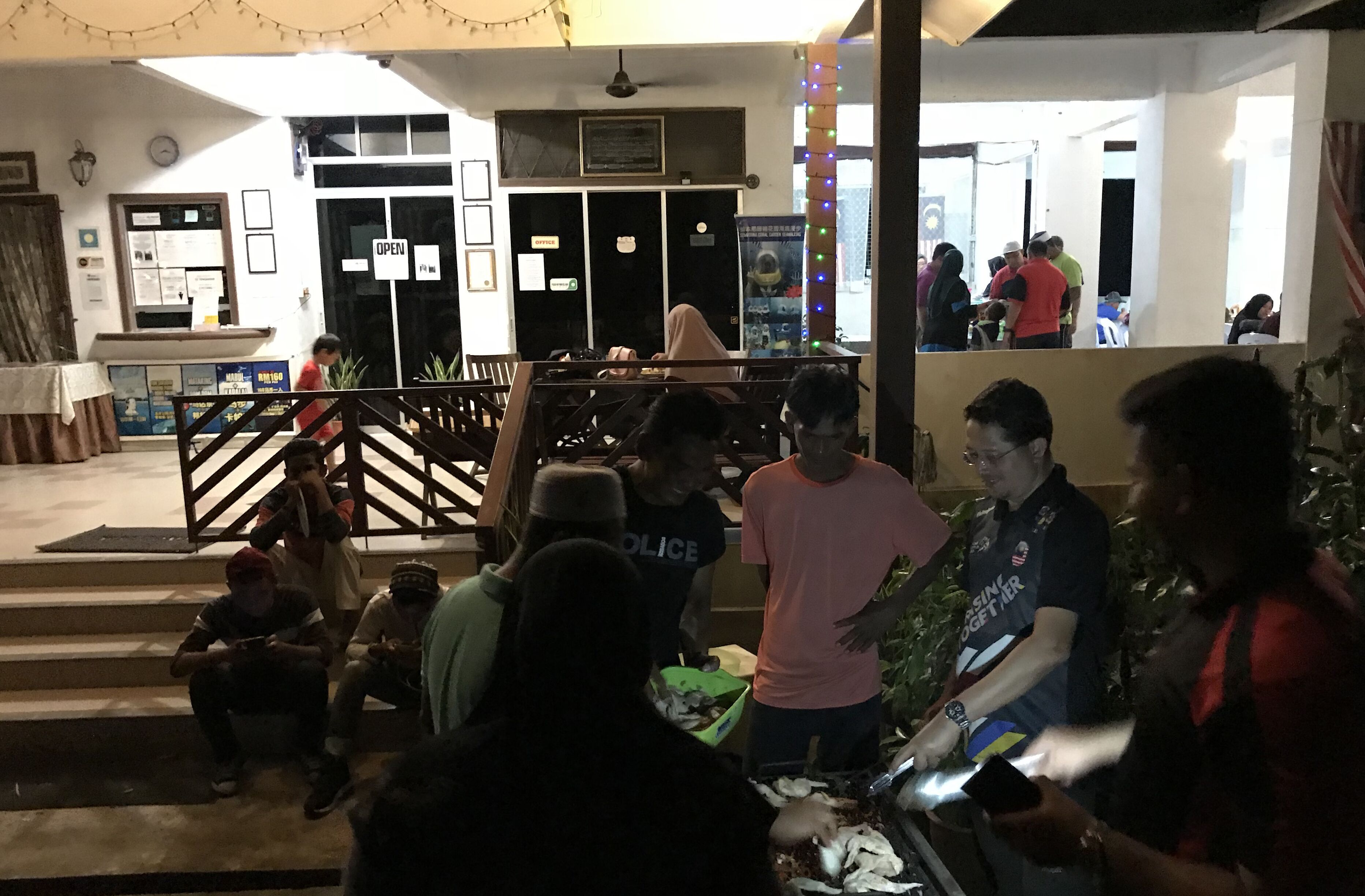 Un hall de hotel con una recepción, una escalera y un grupo de personas reunidas alrededor de una mesa.