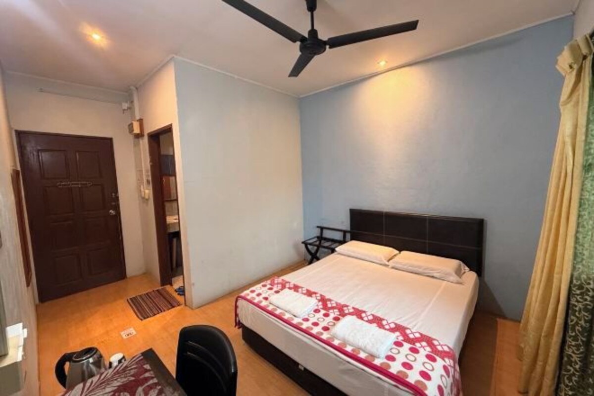 A hotel room with a bed, a desk, a chair, a ceiling fan, and a door.