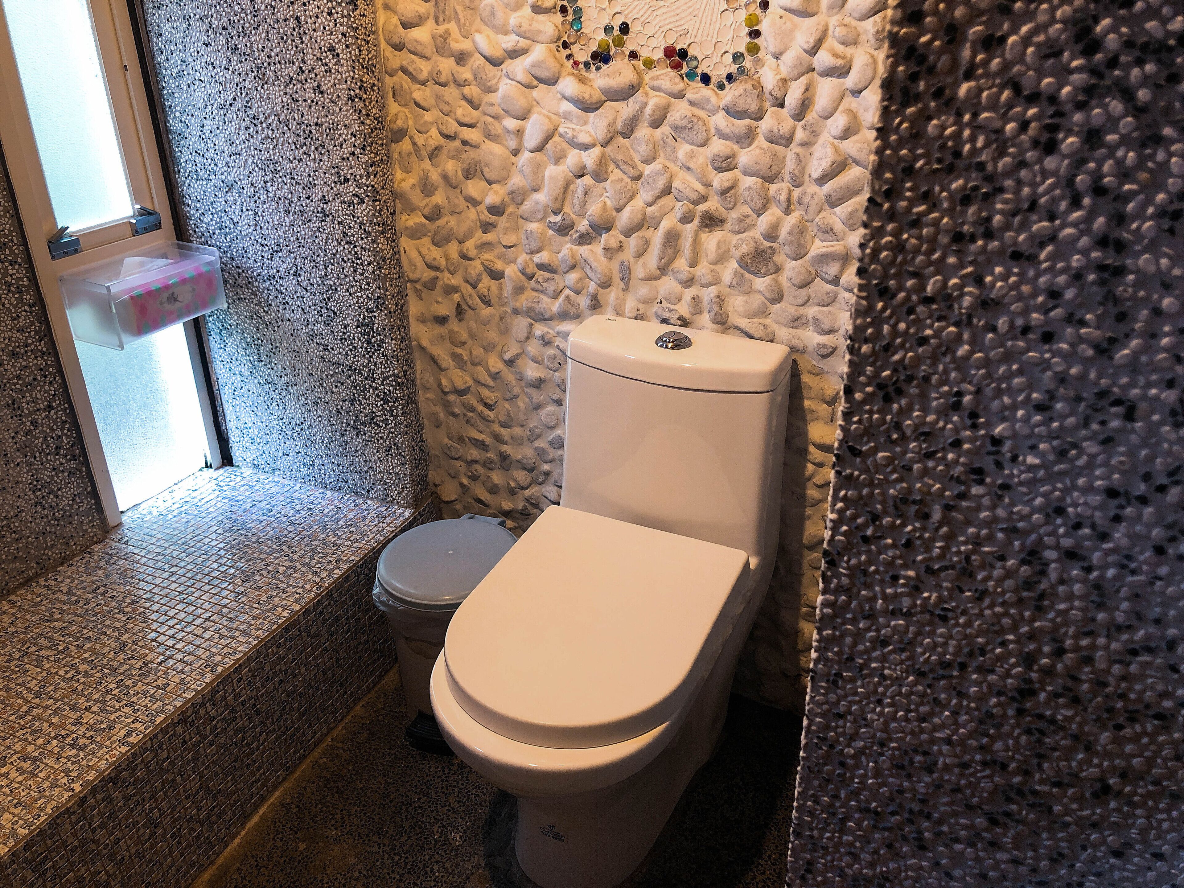 Un baño con pared de piedra, un inodoro y un cubo de basura.