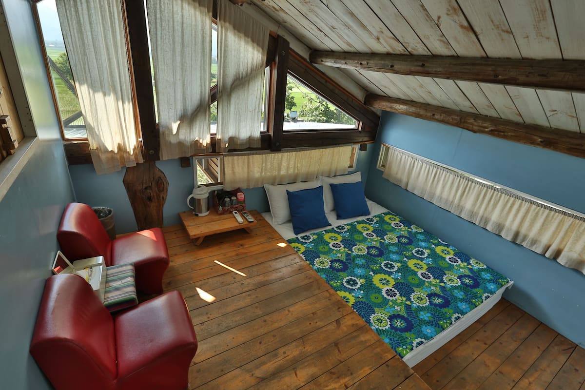 A cozy room with a wooden ceiling, a bed with a colorful blanket, a red sofa, and a small table with a remote control.