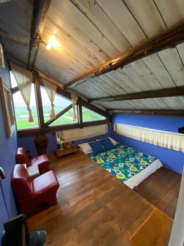 Een kamer met een schuine plafond, houten vloer, een bed met een kleurrijk sprei, een kleine tafel en twee rode stoelen.