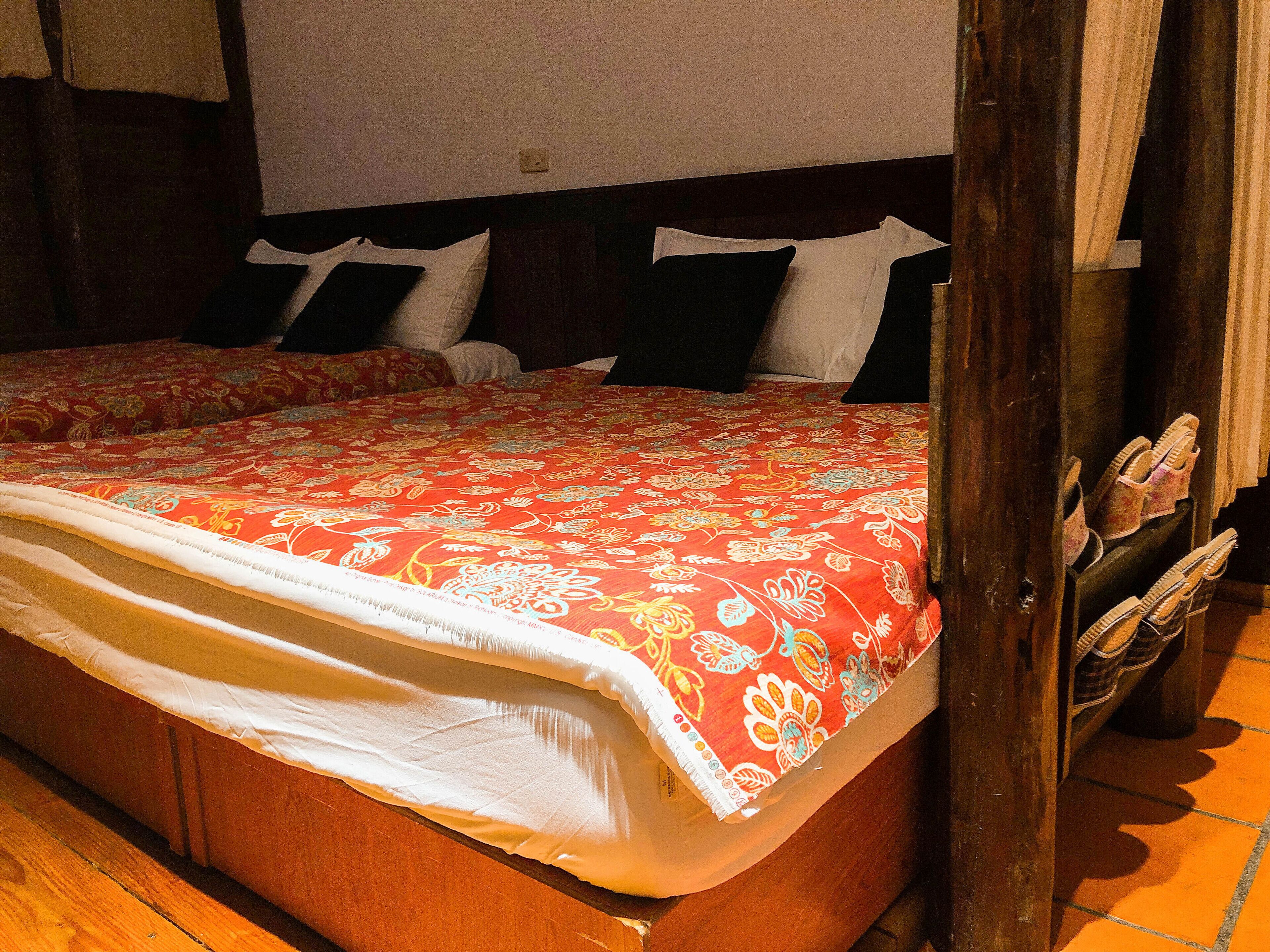 Un lit avec une couvre-lit à motifs floraux, une tête de lit en bois et une paire de chaussons posés sur un petit meuble en bois.
