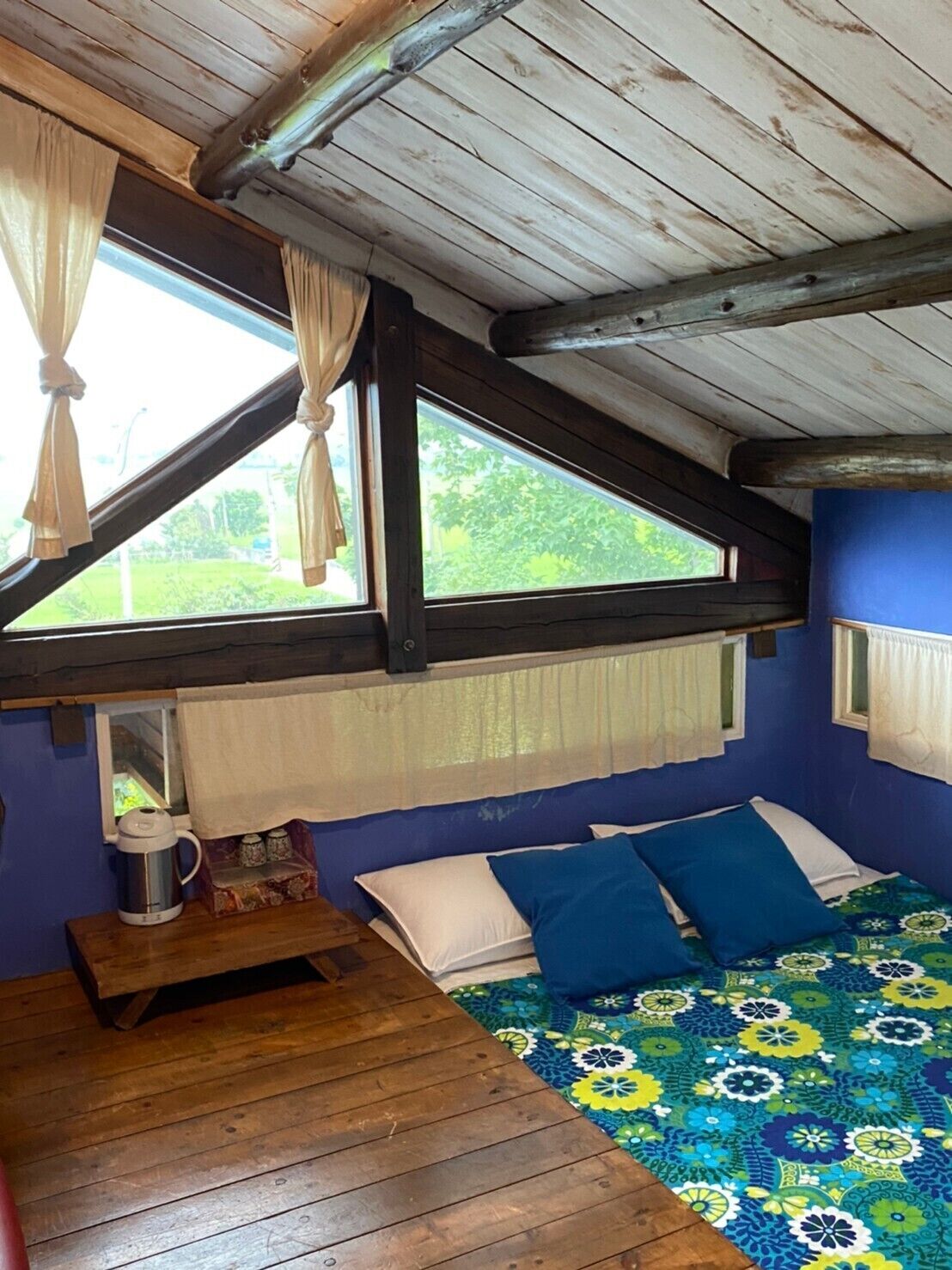 A small, rustic room with a sloped ceiling, a bed with blue and white bedding, a wooden table with a kettle, and a window with curtains.
