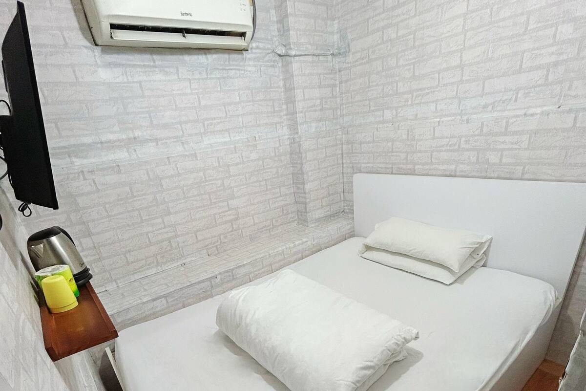 A single bed with white bedding in a room with white brick-patterned walls and a white air conditioning unit mounted above it.