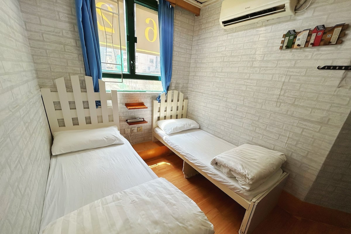 A small, cozy room with two single beds, a wooden headboard, a small shelf, and a window with curtains.