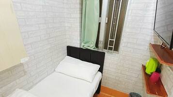 Ein kleines Zimmer mit einem Einzelbett, einer weißen Backsteinwand, einem Fenster mit grünem Vorhang, einem Holzboden, einer kleinen Mülltonne und einem Fernseher auf einem Holzregal.