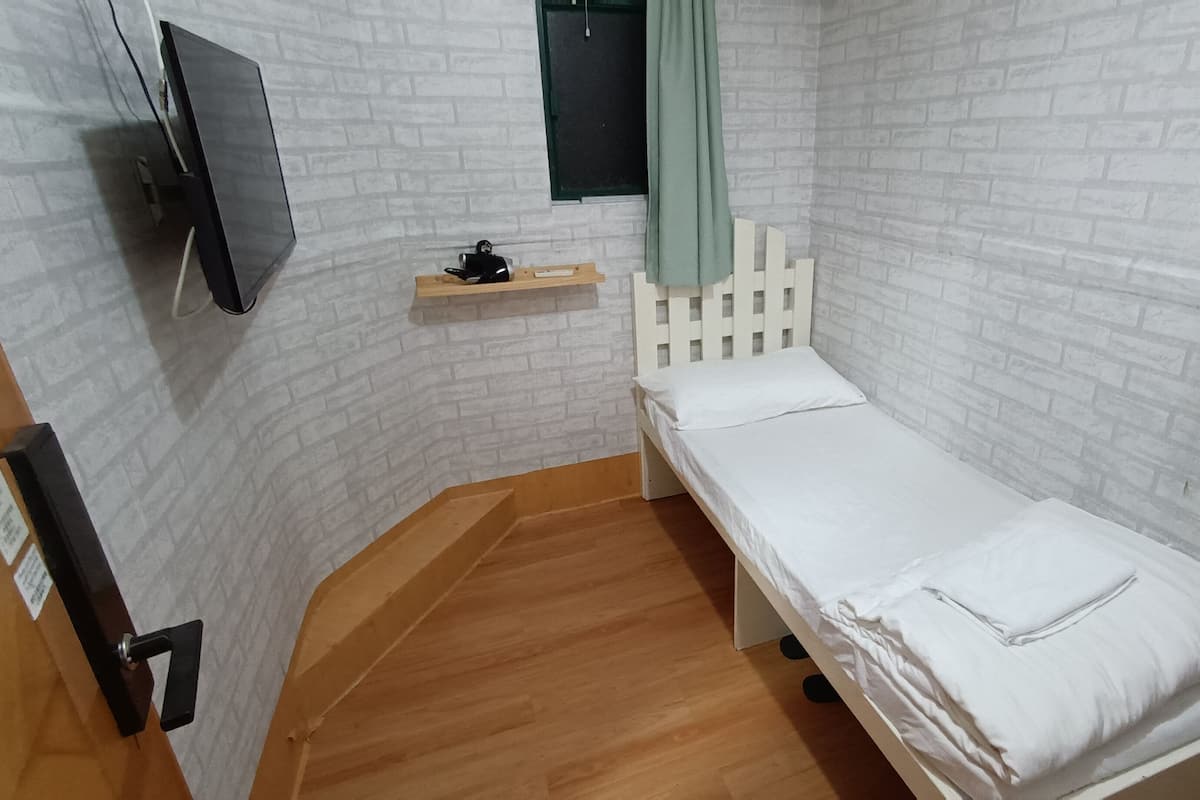 A small, single-bed room with white brick walls, a wooden floor, a TV mounted on the wall, and a small shelf with a green curtain.