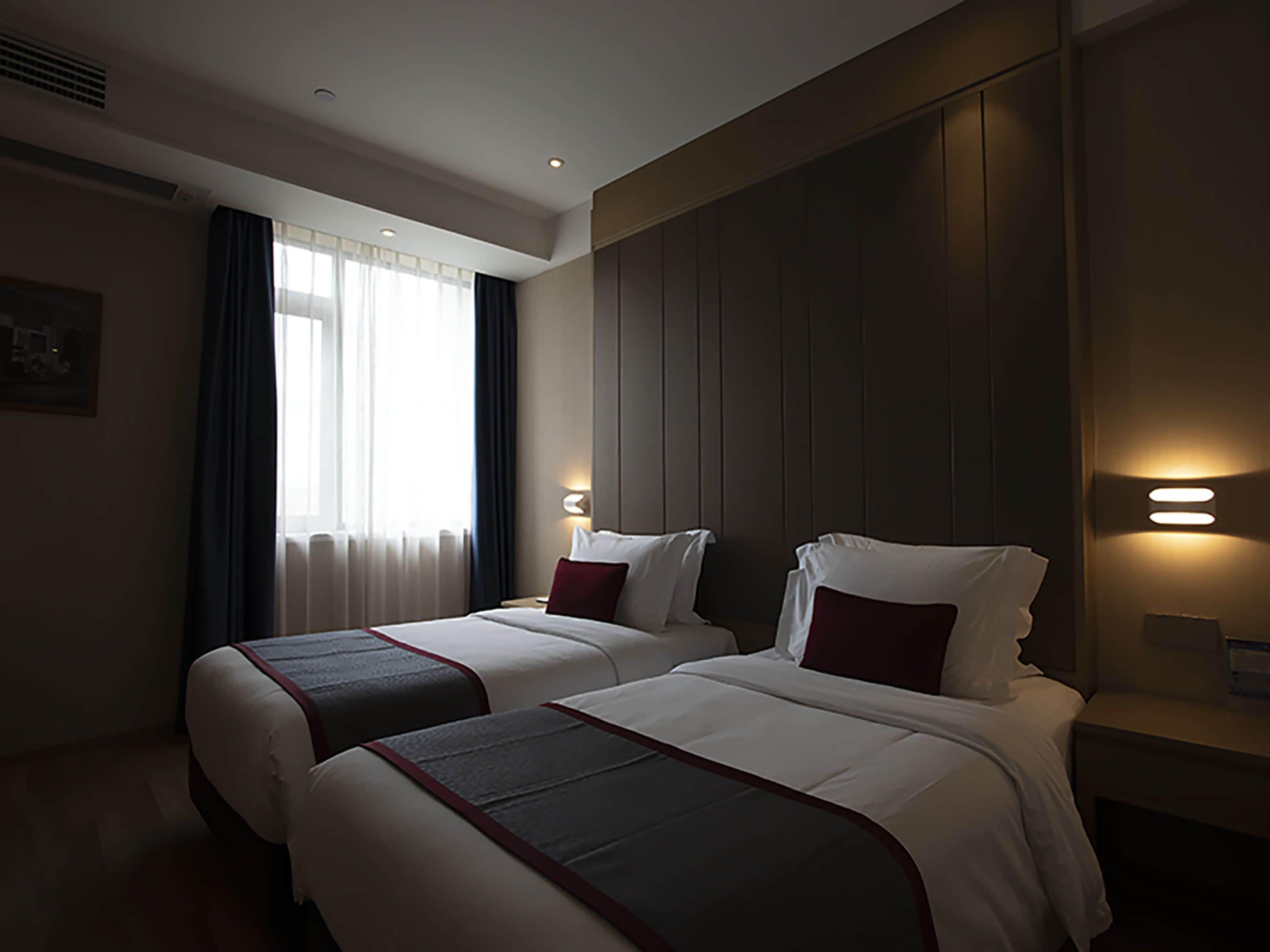 A hotel room with two beds, a large window with curtains, a wall-mounted light, and a painting on the wall.