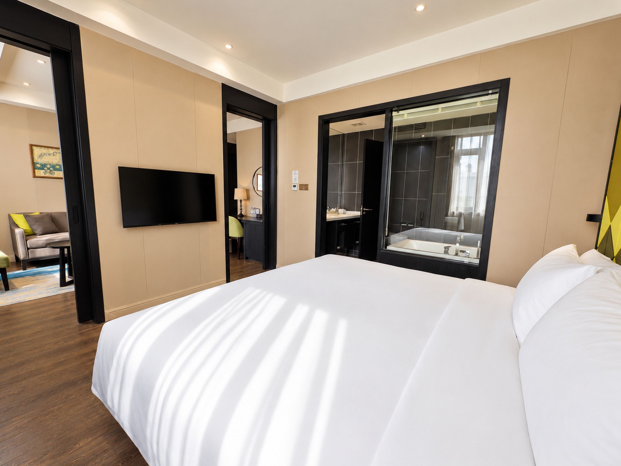 A hotel room with a large bed, a television mounted on the wall, a bathroom with a mirror and sink, and a seating area with a sofa and chair.