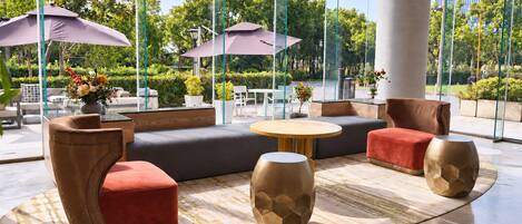 A modern outdoor seating area with sofas, chairs, and tables, surrounded by large windows offering views of greenery.