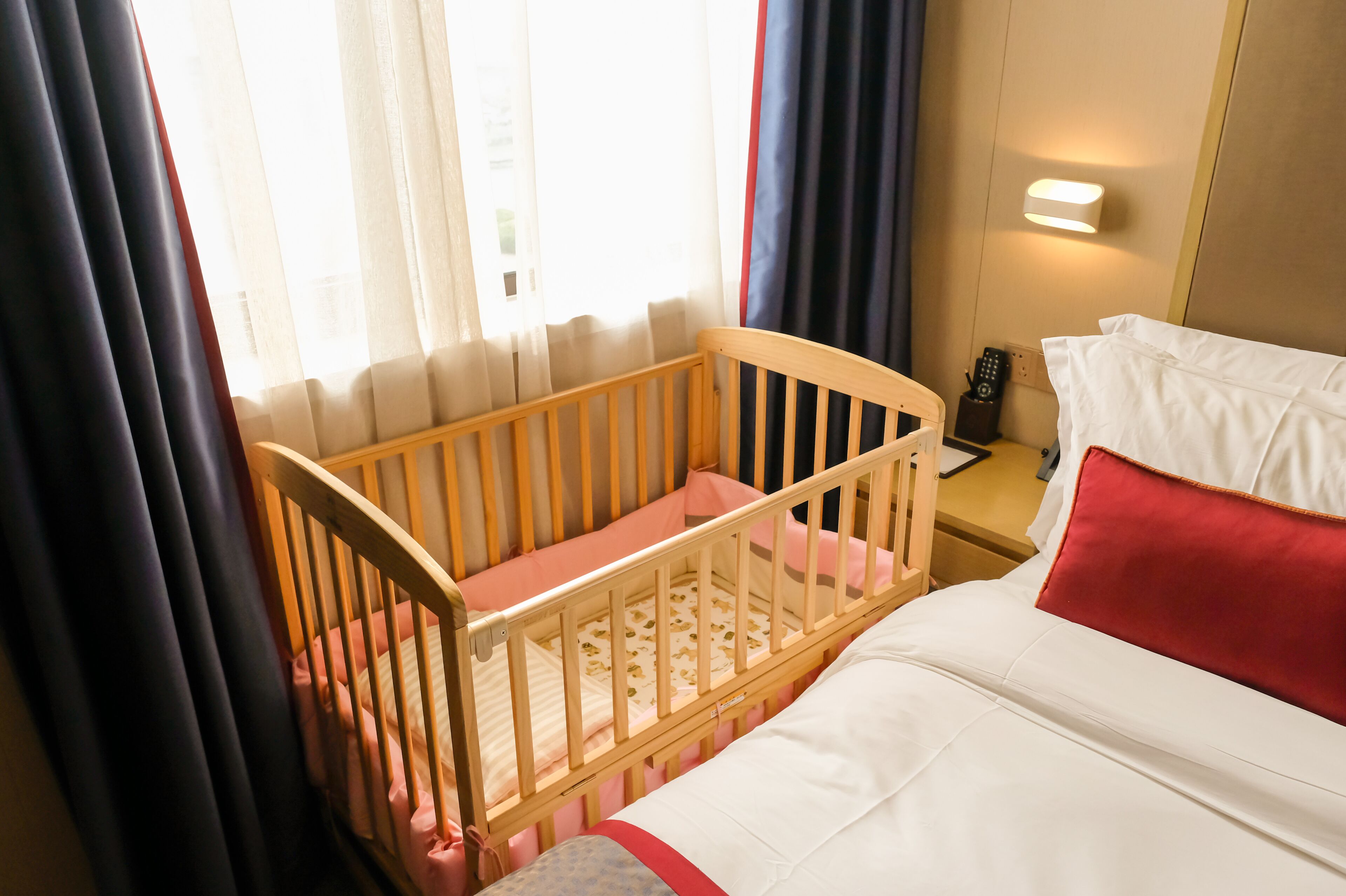 Hotelkamer met een ledikant, een bed met wit en rode beddengoed, een nachtkastje met een afstandsbediening en een aan de muur bevestigde lamp.