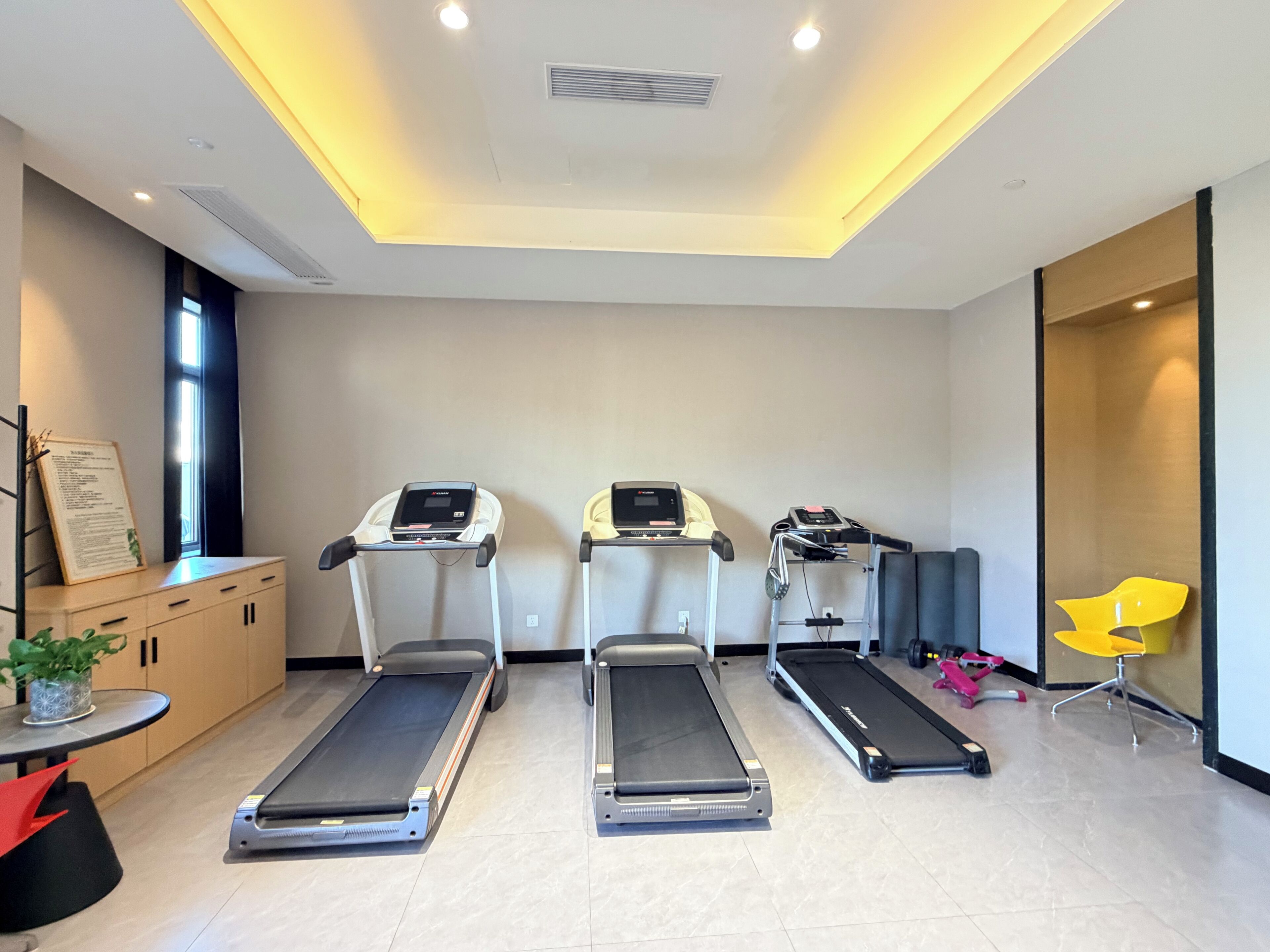 A modern fitness room with three treadmills, a yellow chair, and a small table with a plant.