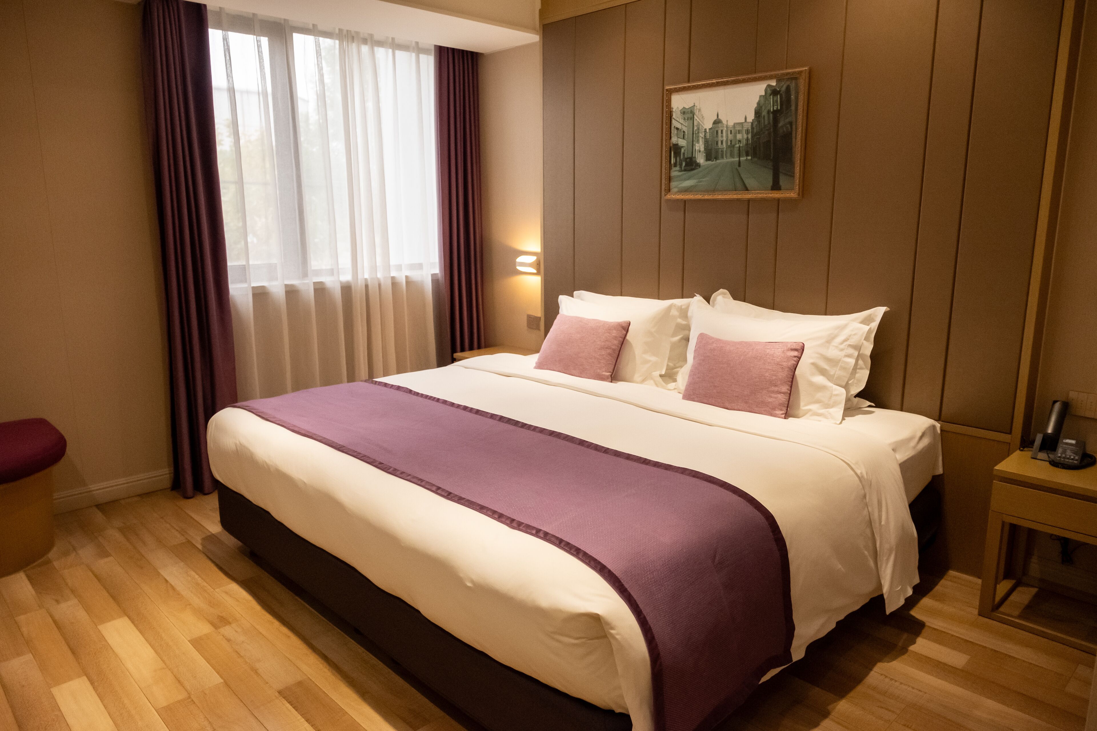 A hotel room with a large bed, a nightstand, a window with curtains, and a painting on the wall.