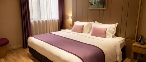 A hotel room with a large bed, a nightstand, a window with curtains, and a painting on the wall.