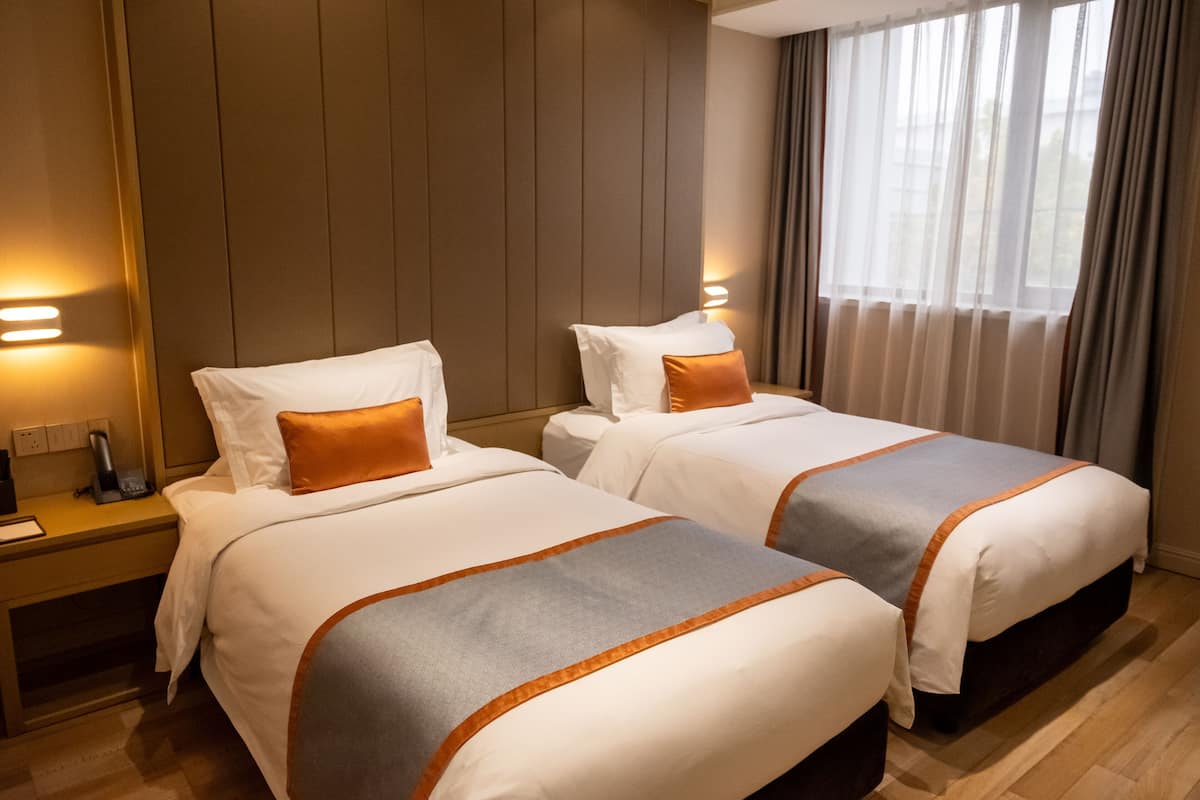 A hotel room with two beds, each with white bedding and orange accents, a wooden headboard, a bedside table with a phone, and a window with curtains.