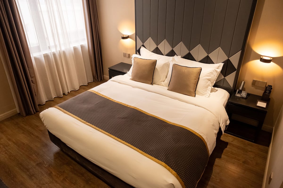 A hotel room with a large bed, two bedside tables, a headboard with a geometric pattern, and a window with curtains.