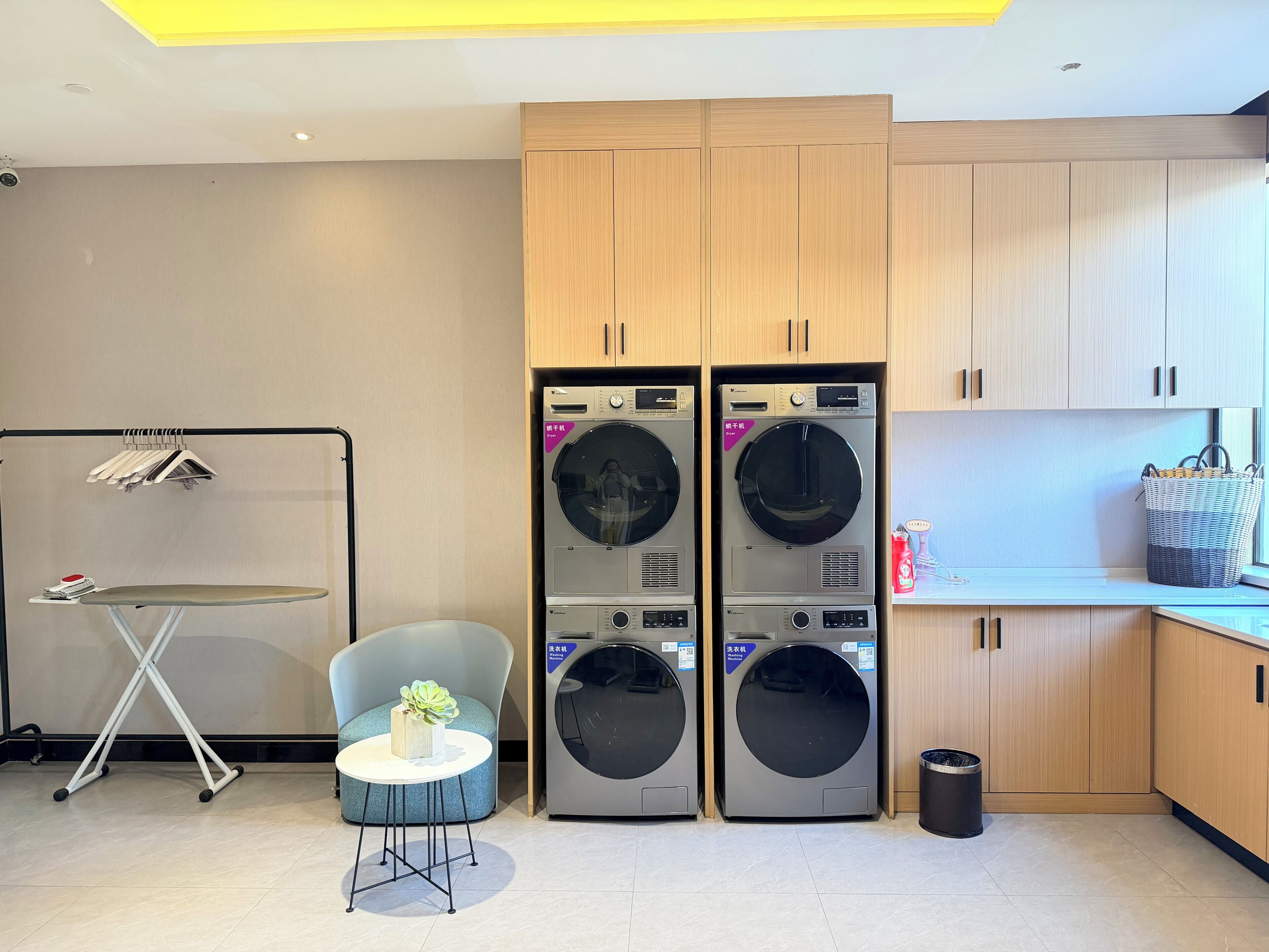 A laundry room with two stacked washing machines, a drying rack, a small table with a plant, and a trash bin.