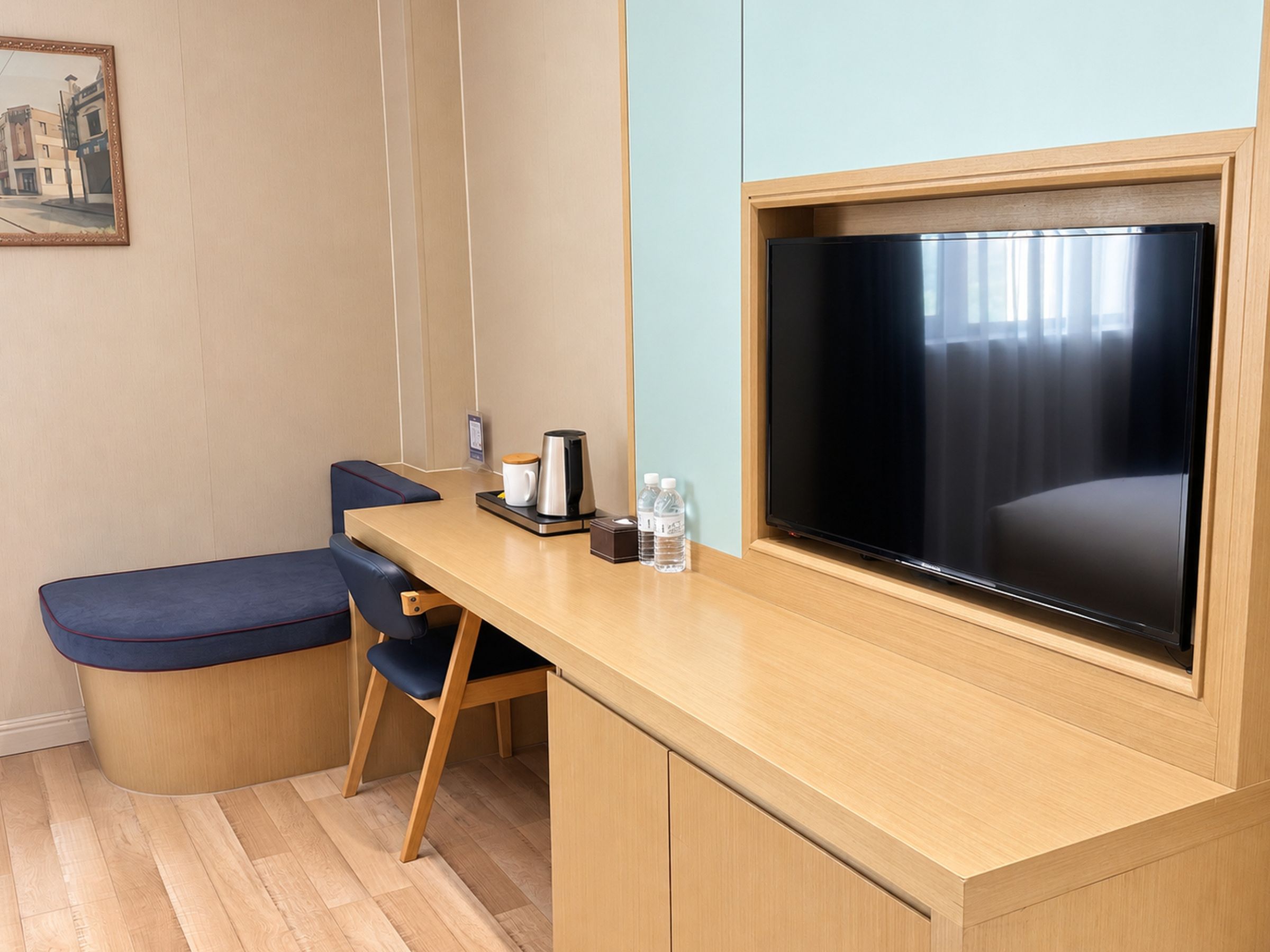 A hotel room with a wooden desk, a television, a cushioned bench, and a painting on the wall.