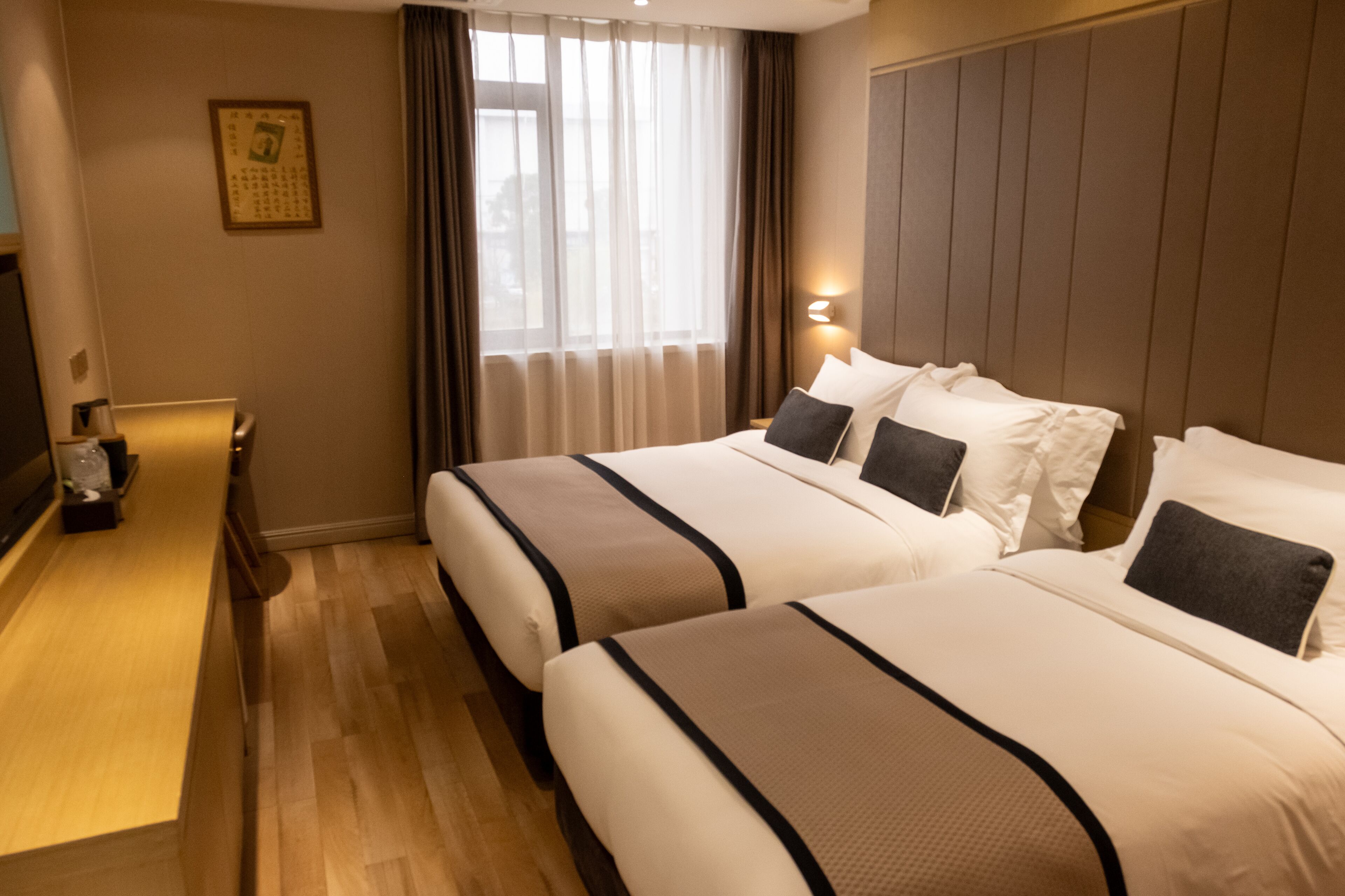 A hotel room with two beds, a wooden floor, a nightstand, a lamp, and a window with curtains.