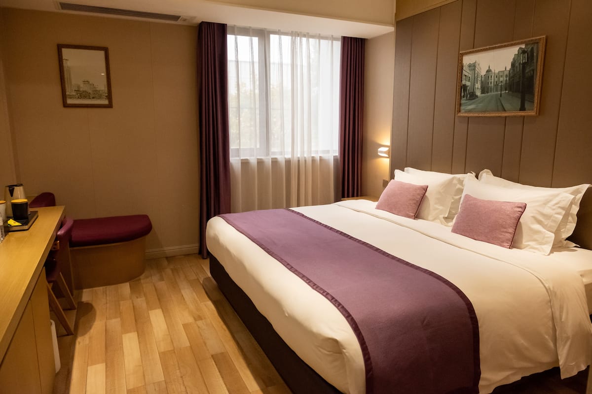 A hotel room with a large bed, a desk with a chair, a bench, a window with curtains, and a framed picture on the wall.