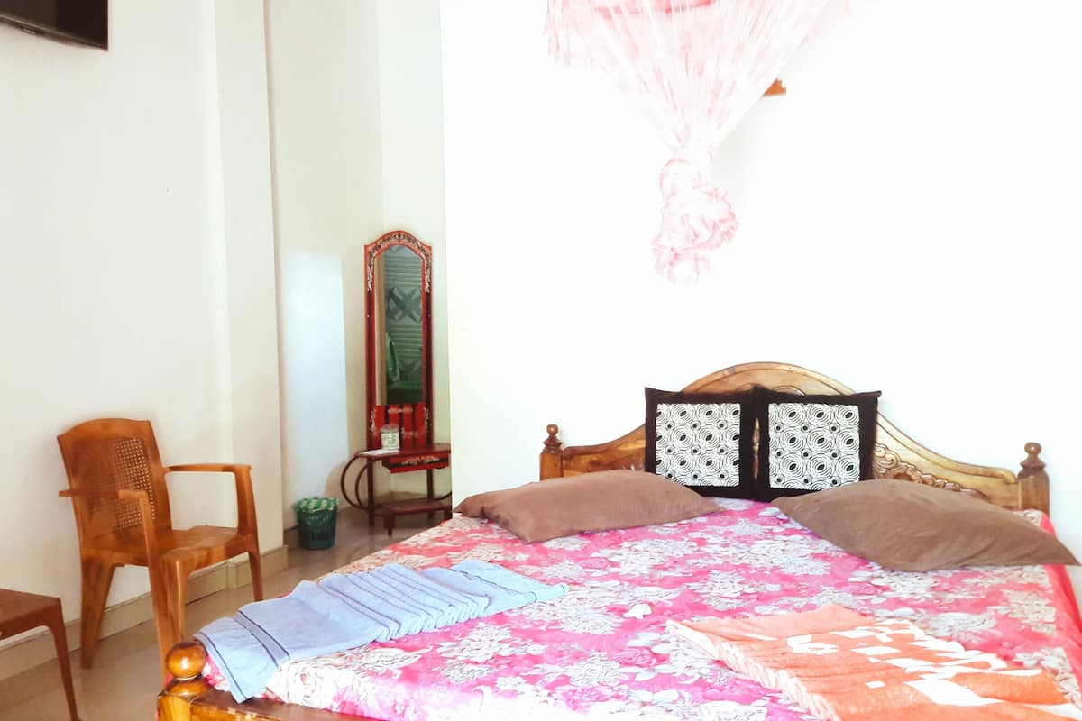 A bedroom with a bed, a chair, a small table, a mirror, and a television on the wall.