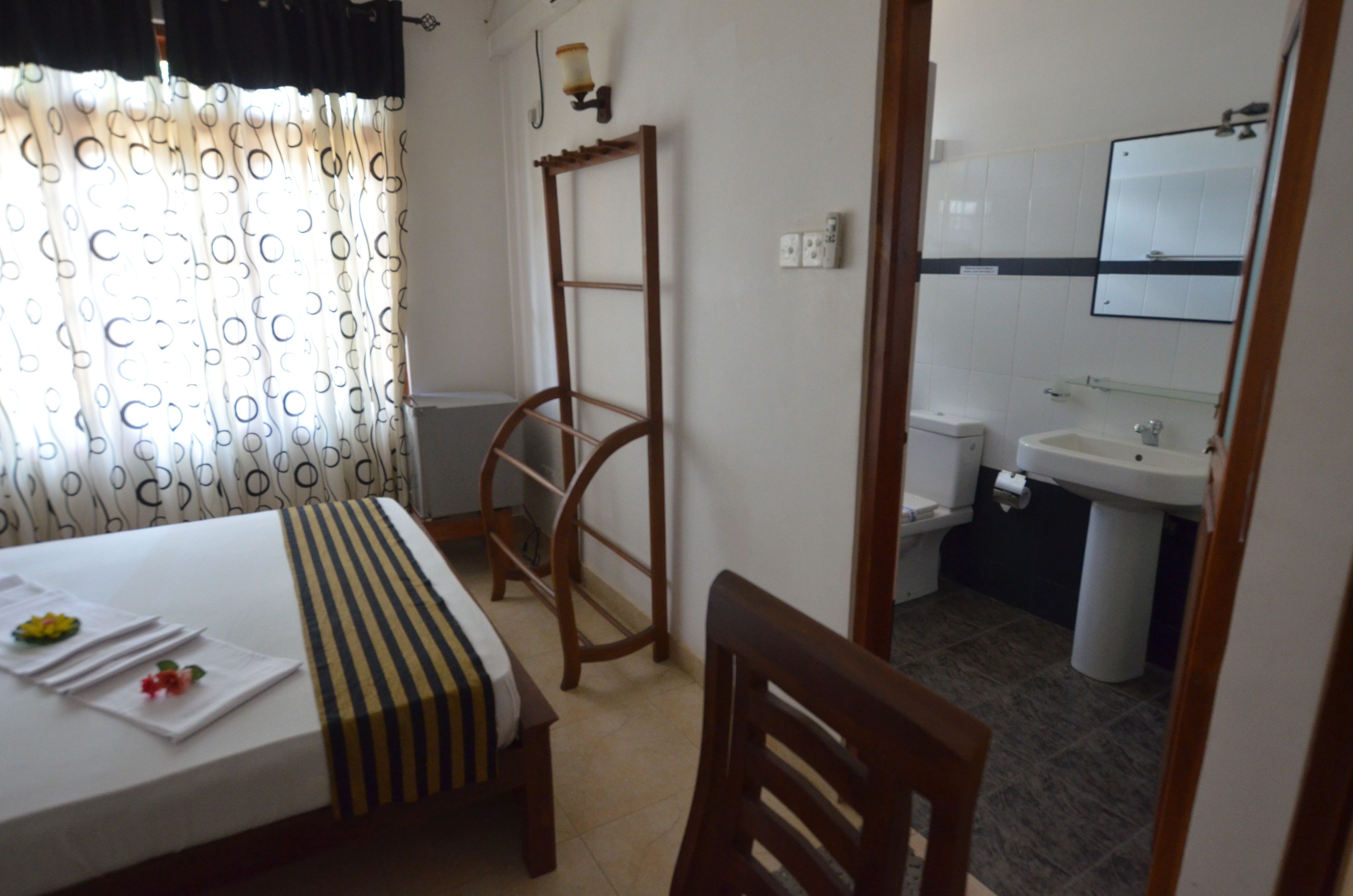 Habitación de hotel con cama, una silla de madera, una mesita con una toalla y flores, una ventana con cortinas estampadas, una escalera de madera, un lavamanos y un espejo.