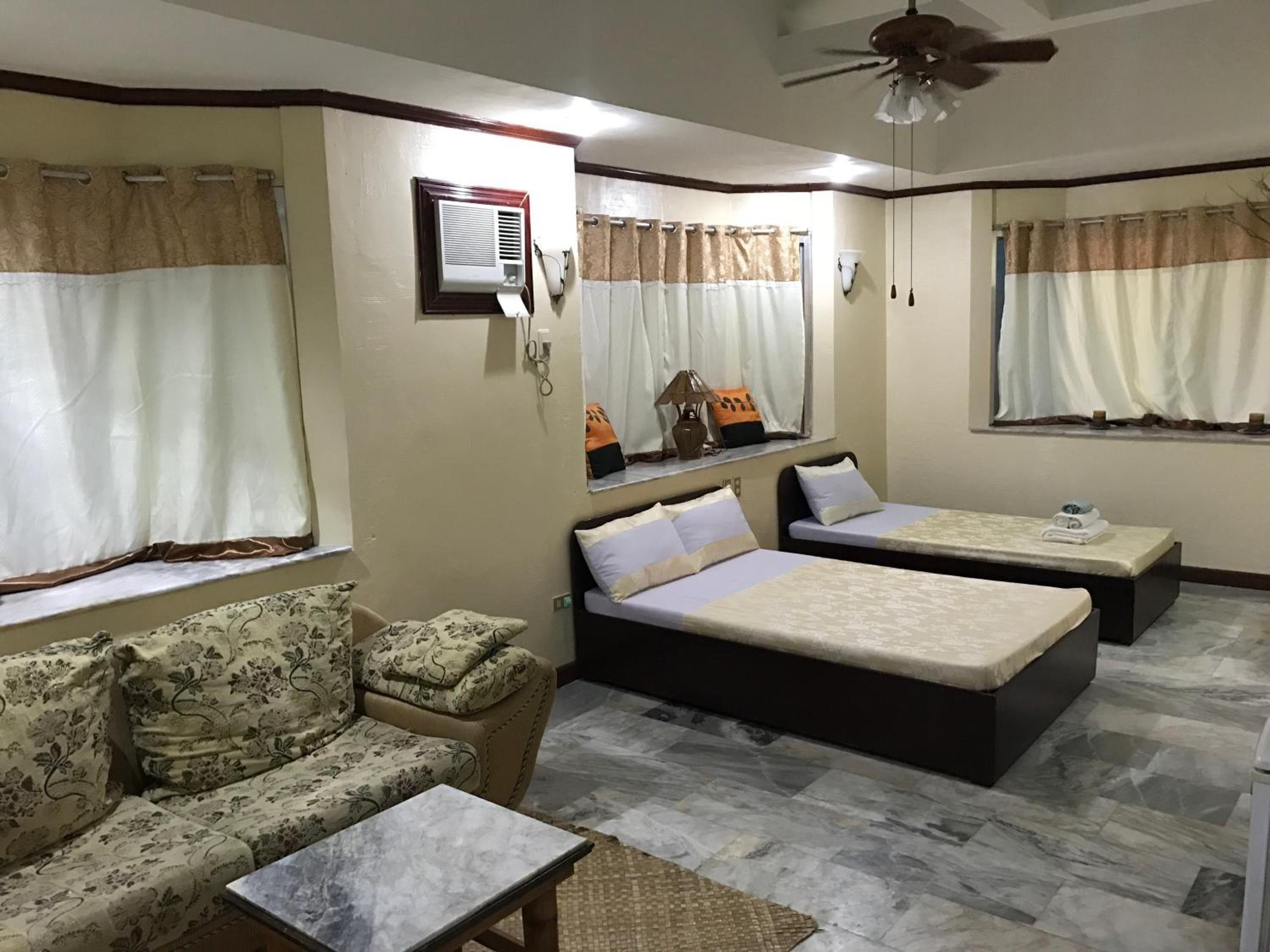 A hotel room with two beds, a sofa, a small table, and a ceiling fan.
