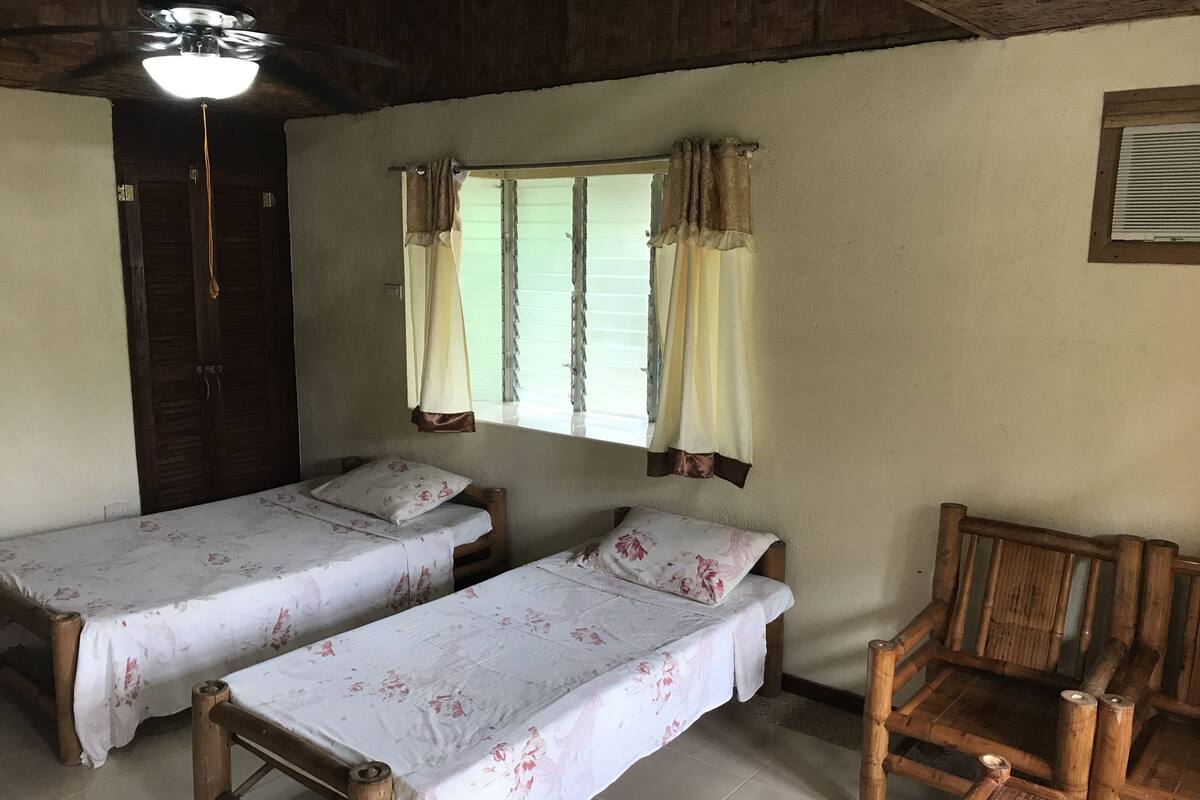 Una habitación con dos camas individuales, una silla de madera, una ventana con cortinas y un ventilador de techo.