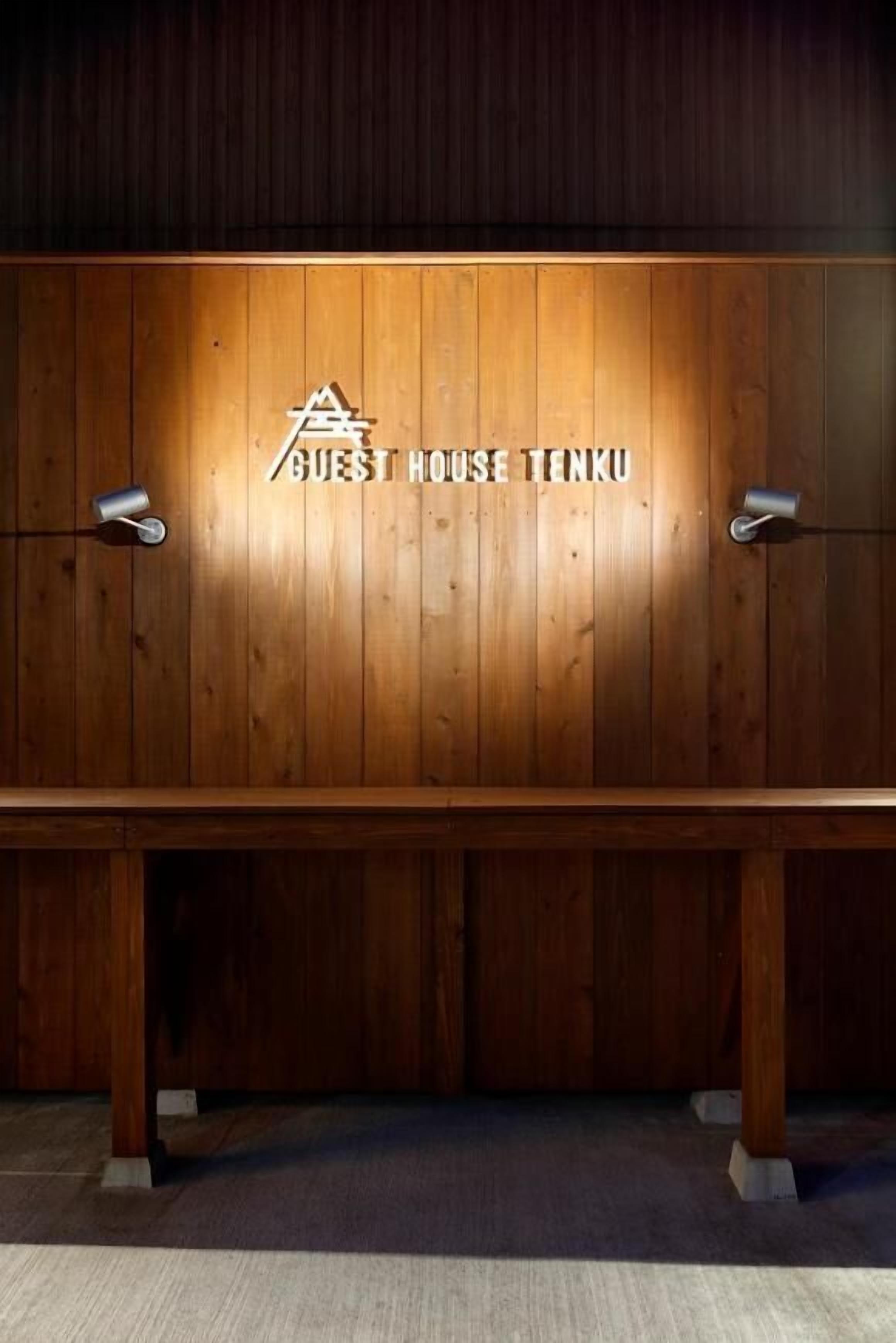 Pared de madera con el texto "GUEST HOUSE TENKU" y dos lámparas a cada lado.