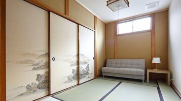 Una habitación japonesa tradicional con suelo de tatami, un biombos shoji con arte paisajístico, un sofá y una pequeña mesa con lámpara.