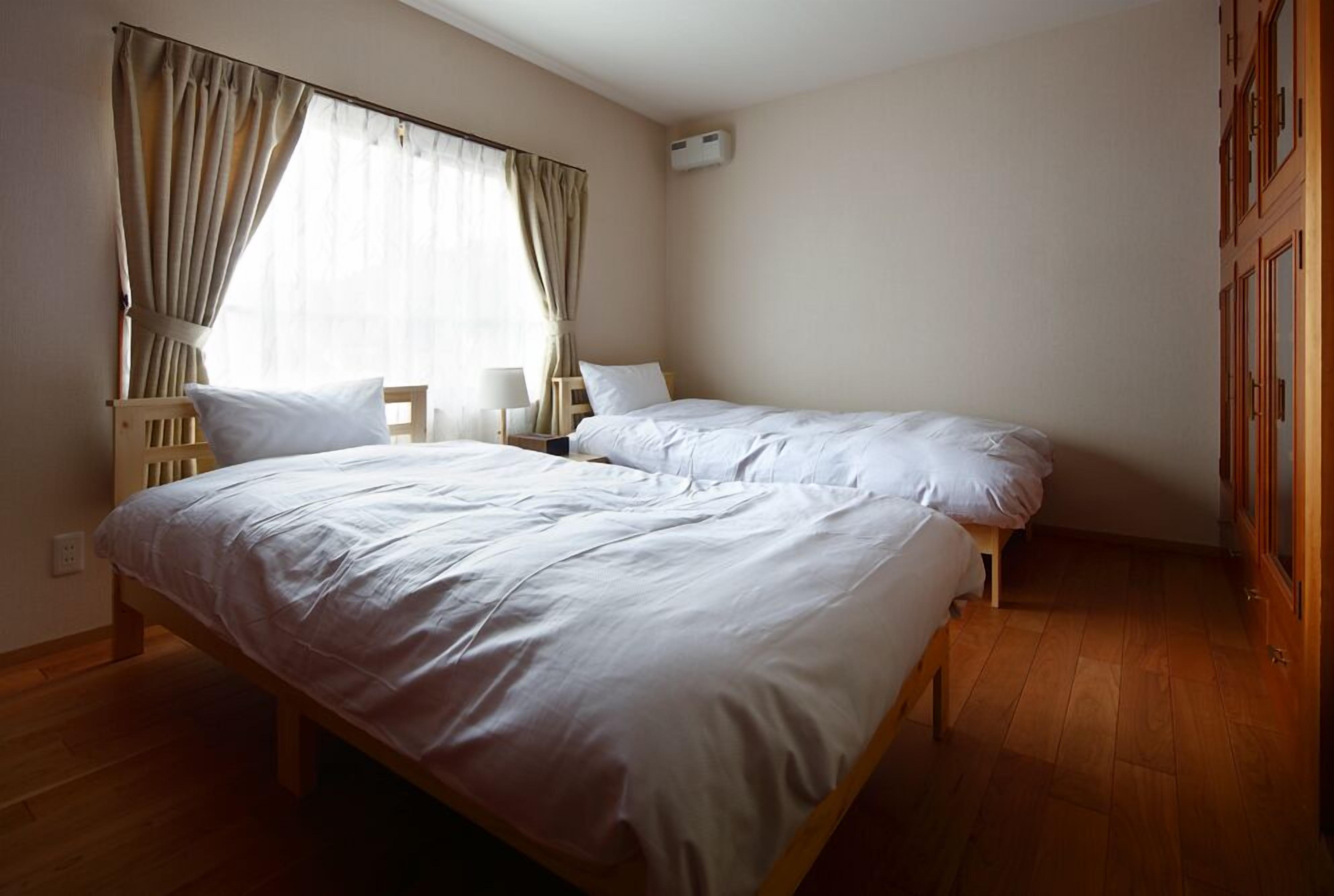 A room with two single beds, a wooden floor, a window with curtains, a bedside table with a lamp, and a wardrobe.