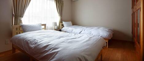 Une chambre avec deux lits simples, un plancher en bois, une fenêtre avec des rideaux, une table de chevet avec une lampe et une armoire.