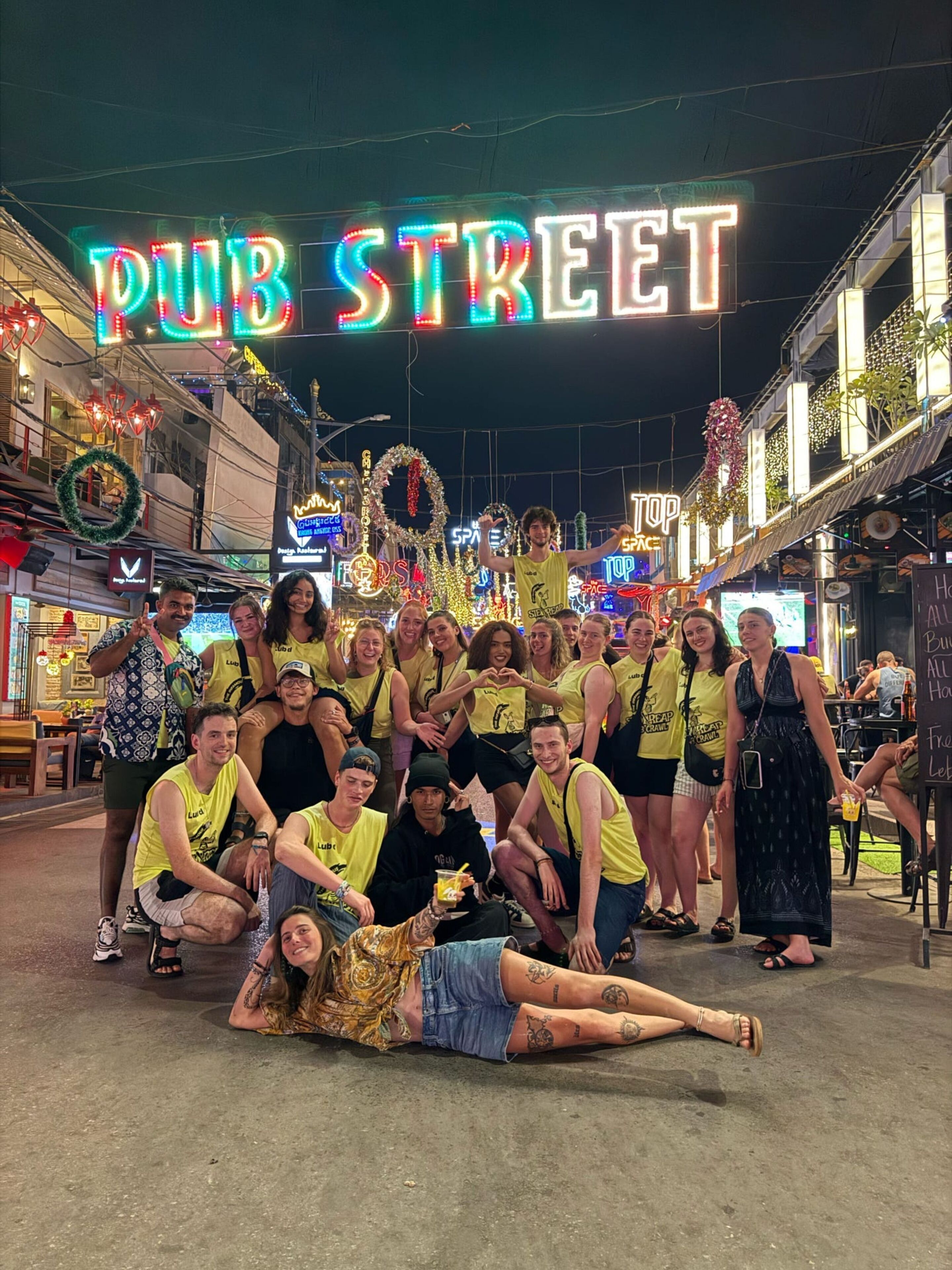 Un groupe de personnes posant devant une enseigne au néon sur laquelle on peut lire « PUB STREET », avec divers commerces et décorations en arrière-plan.