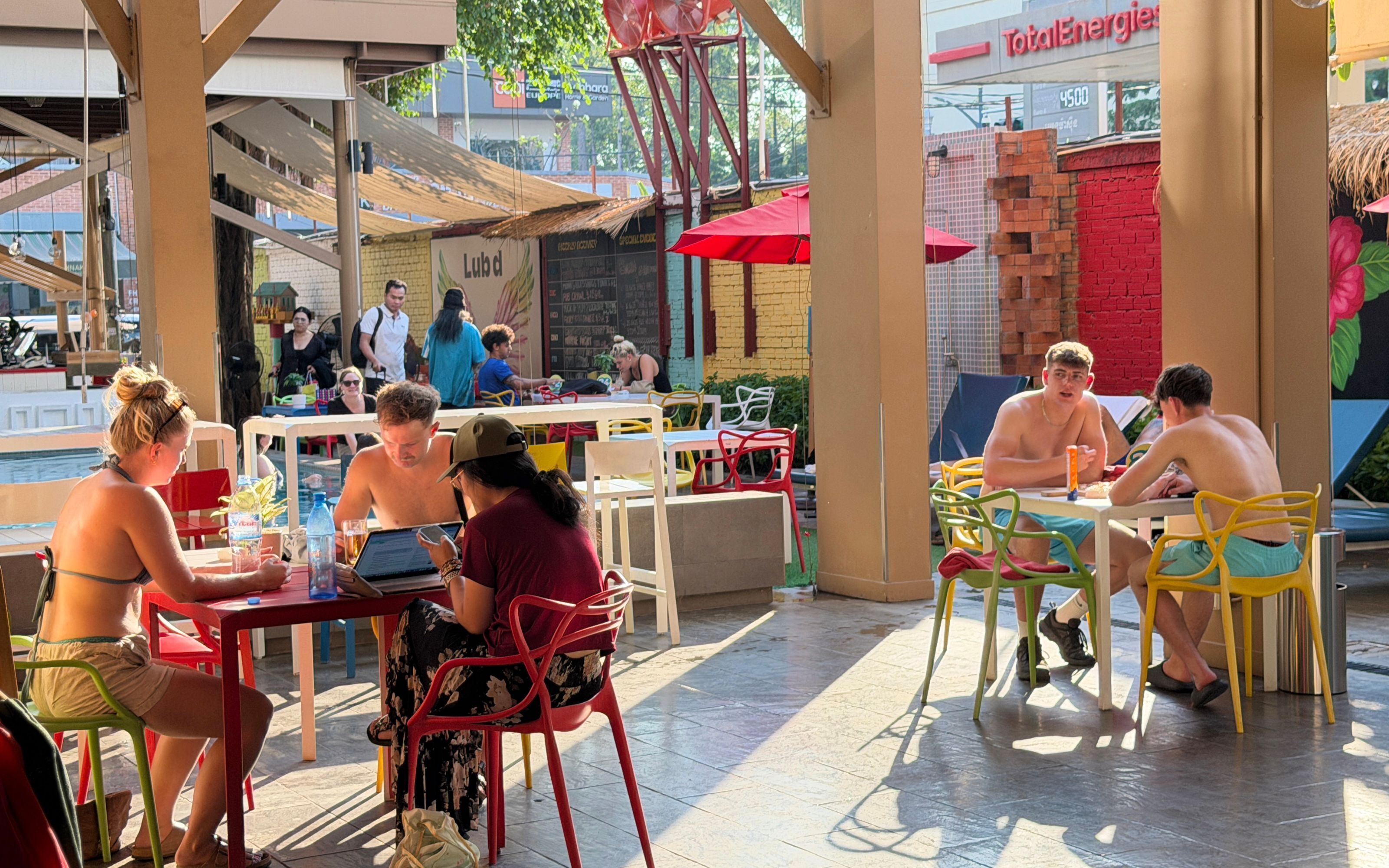 An outdoor dining area with tables and chairs, a pool, and a TotalEnergies sign in the background.