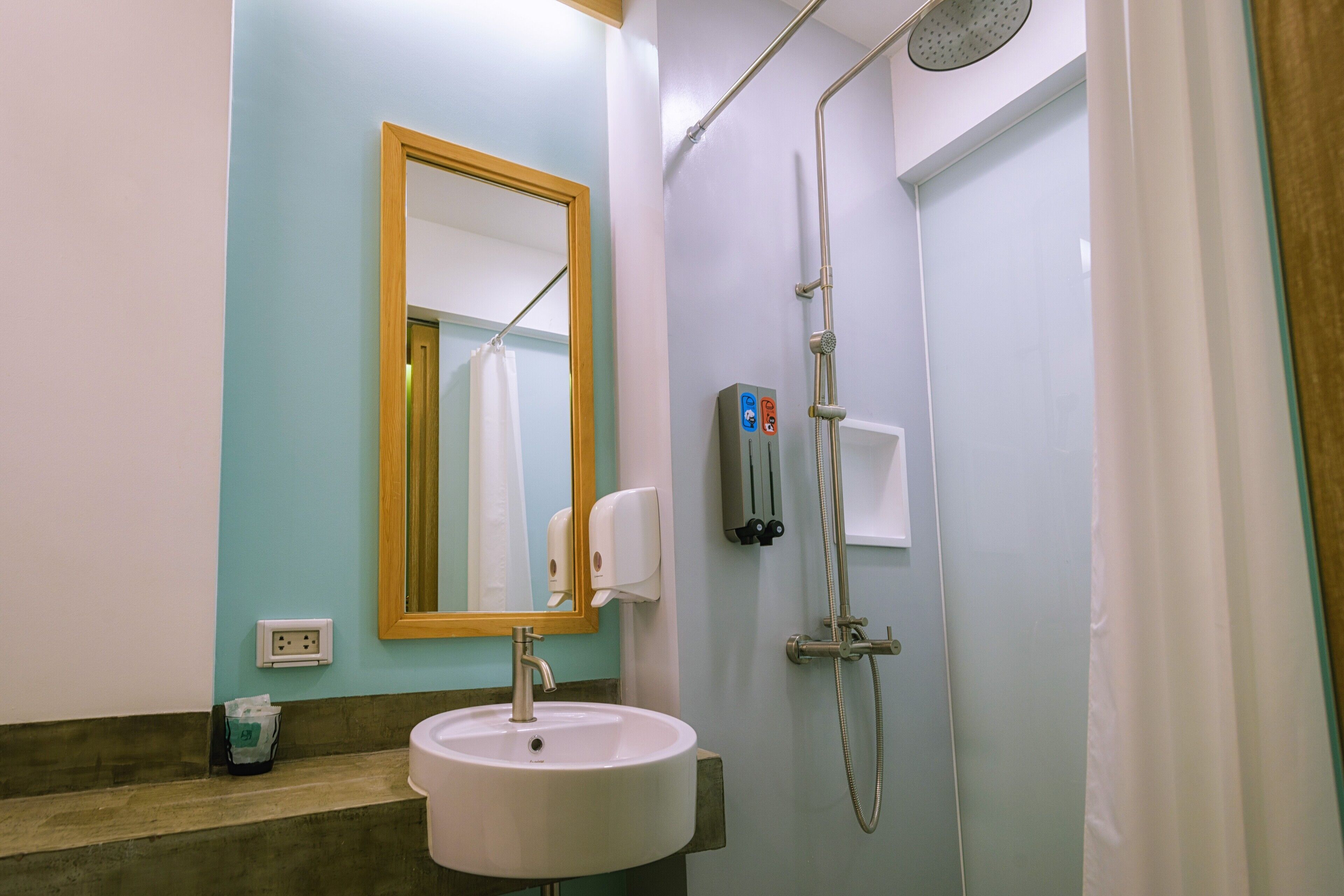 Baño con lavamanos, espejo, ducha y dispensador de jabón.