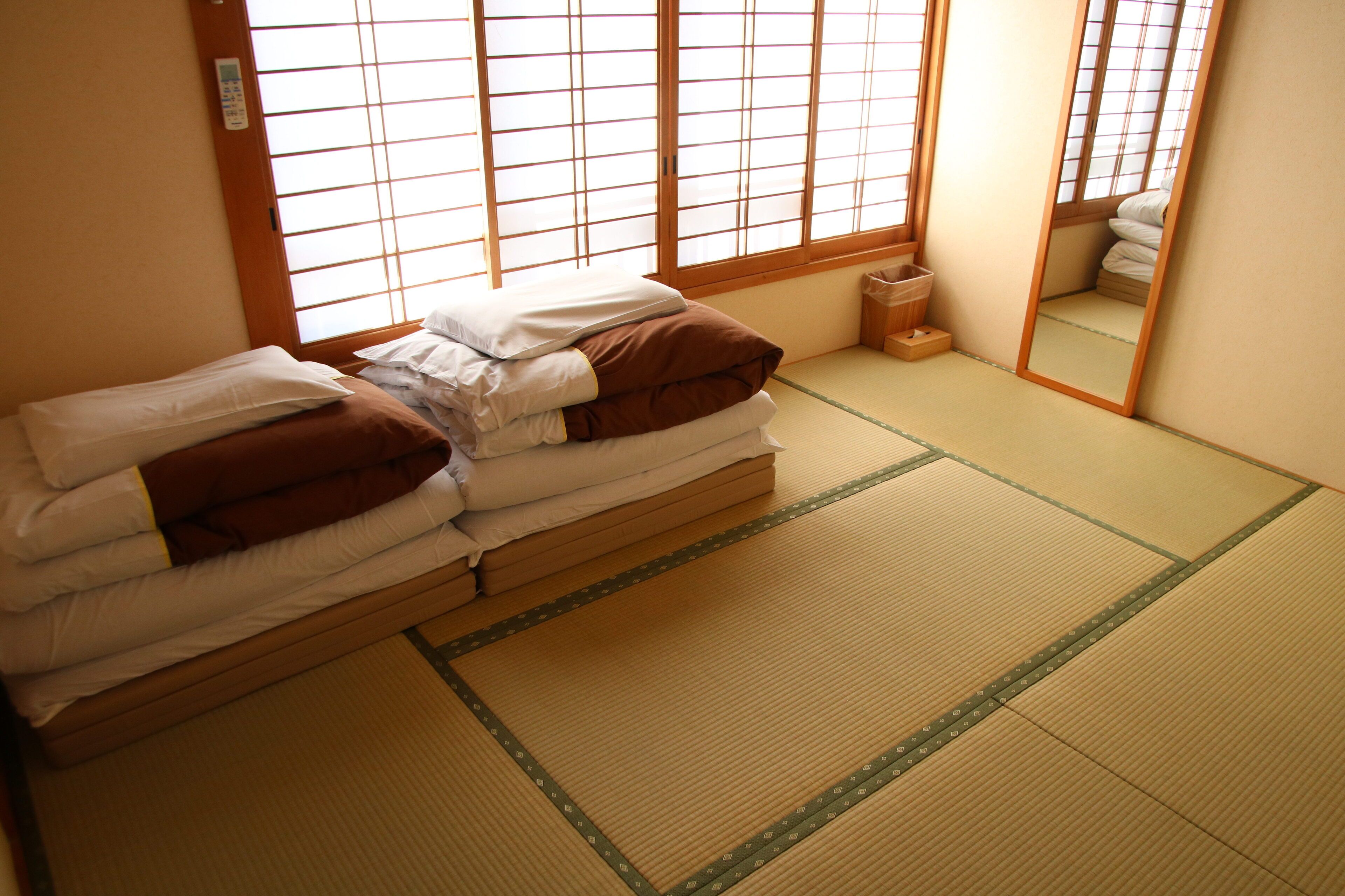 Een traditionele Japanse kamer met tatami vloeren, een laag bed met kussens, een houten kruk en een schuifdeur met uitzicht op het buiten.