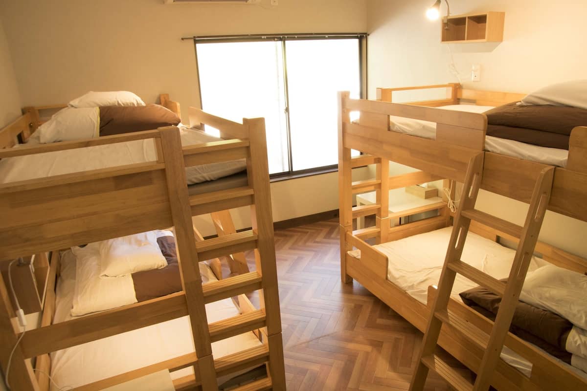 A room with three bunk beds, wooden flooring, a window with blinds, and a small wooden shelf above the beds.
