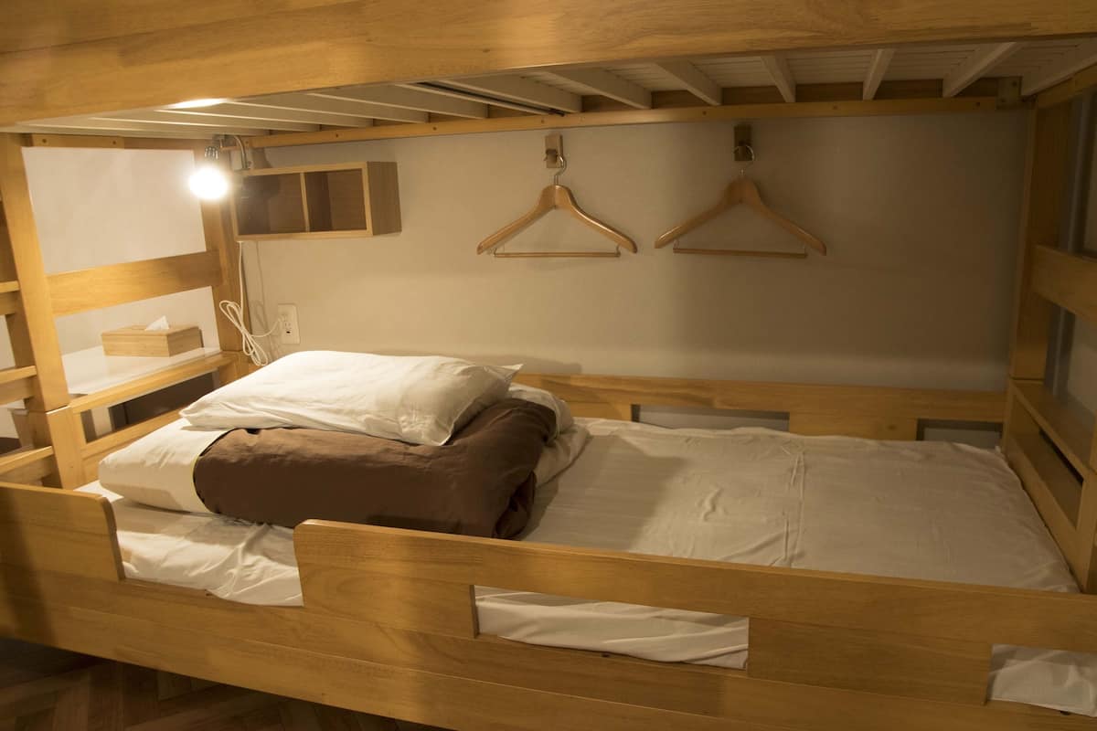 A wooden bunk bed with a single bed on top, a bedside table with a lamp, and two wooden hangers on the wall.