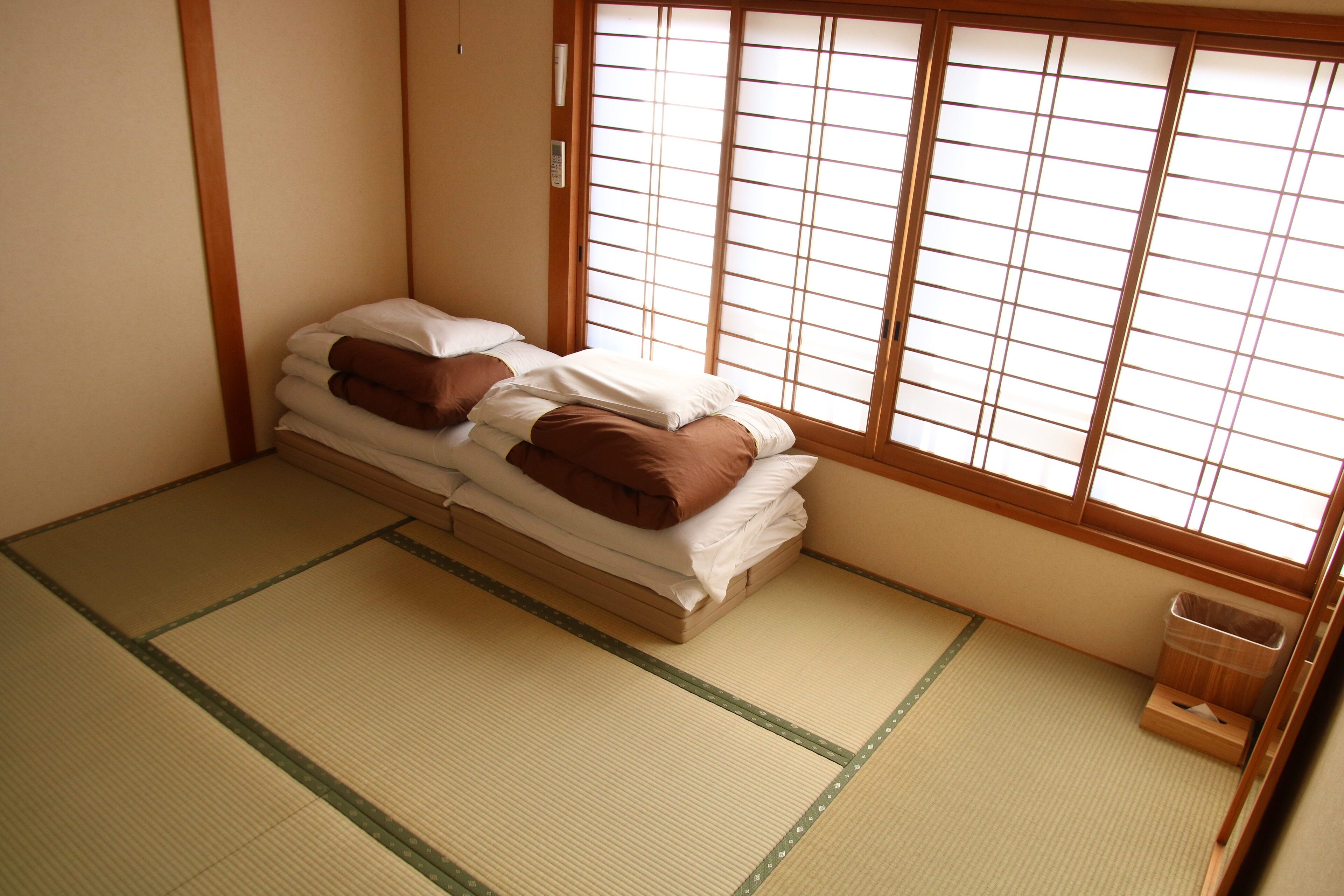 A traditional Japanese room with tatami flooring, a low bed with pillows, and a sliding shoji screen window.