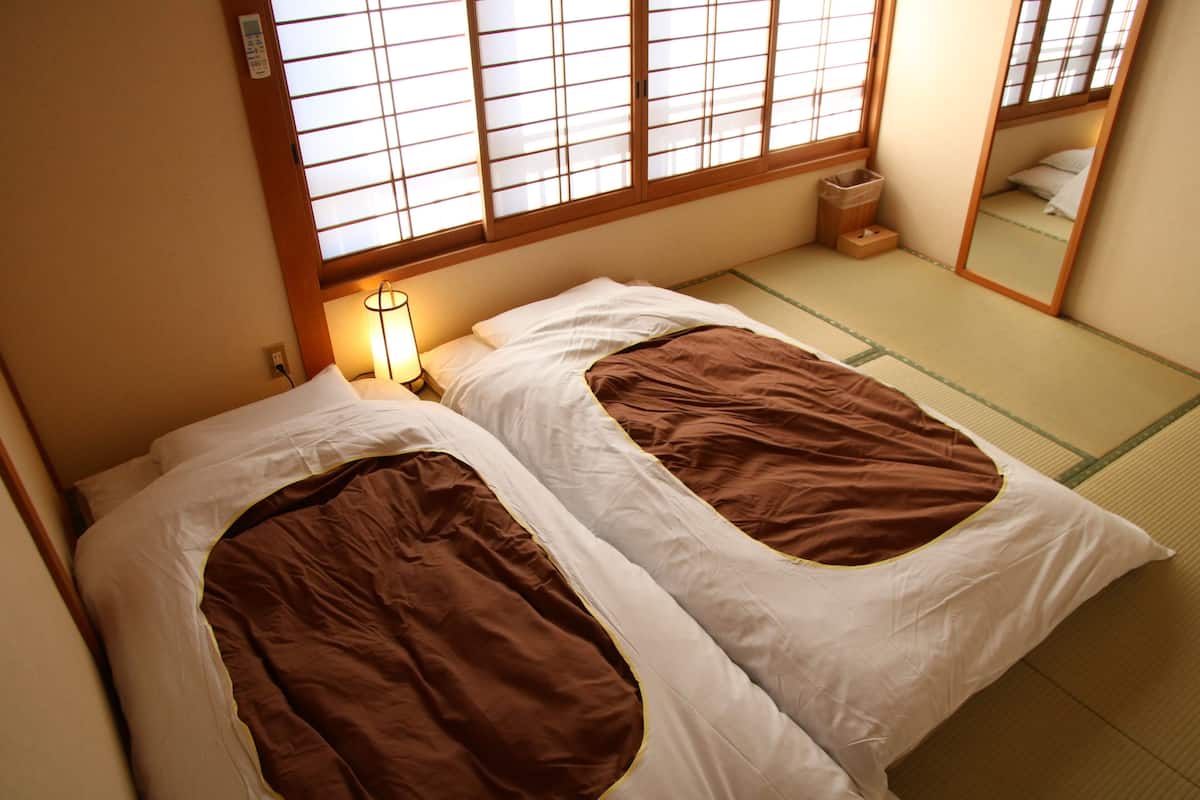 A traditional Japanese room with tatami flooring, two beds with brown bedding, a bedside table with a lamp, and a window with shoji screens.