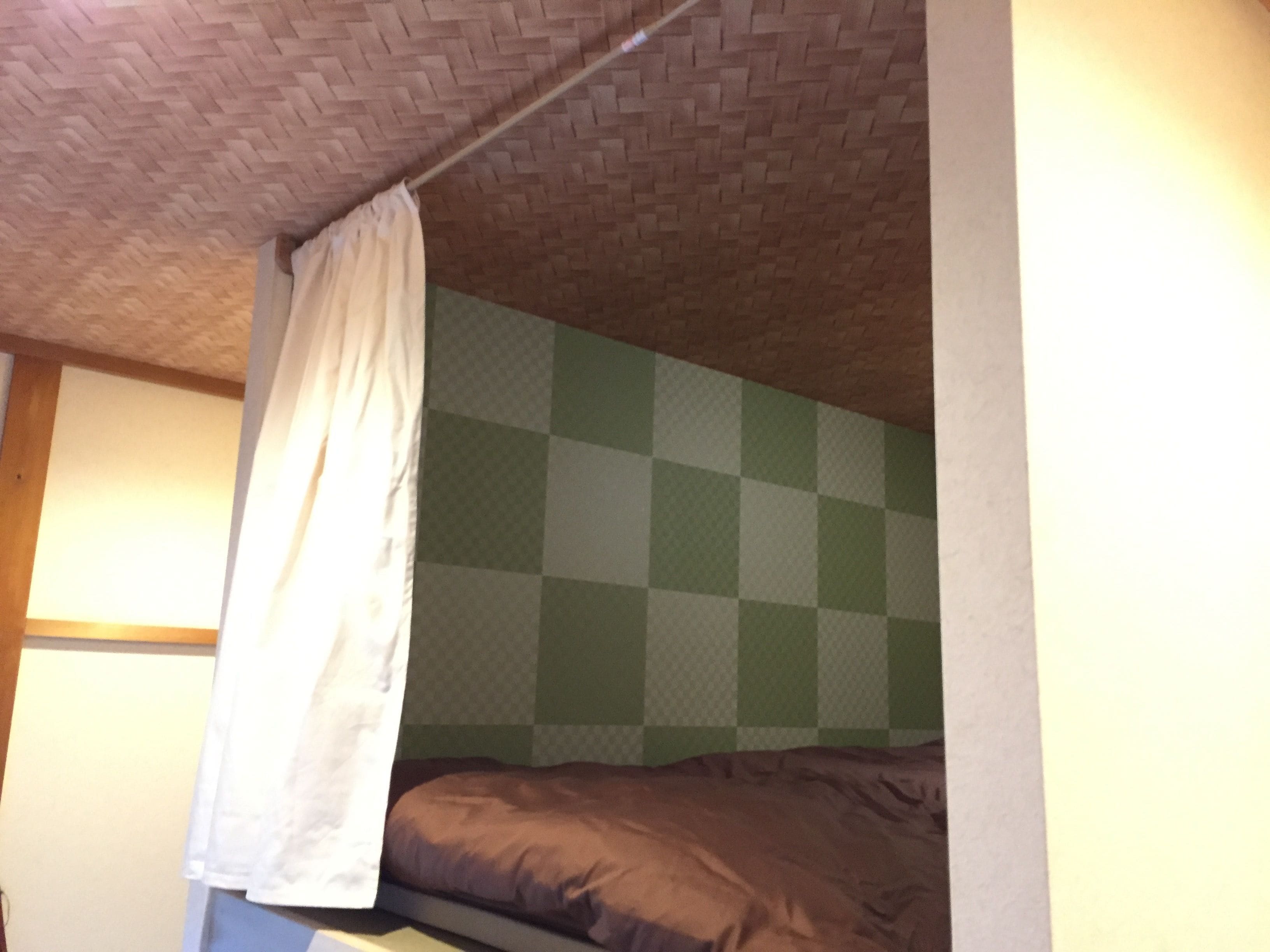 A room with a checkered wall, a bed with a brown mattress, and a white curtain.