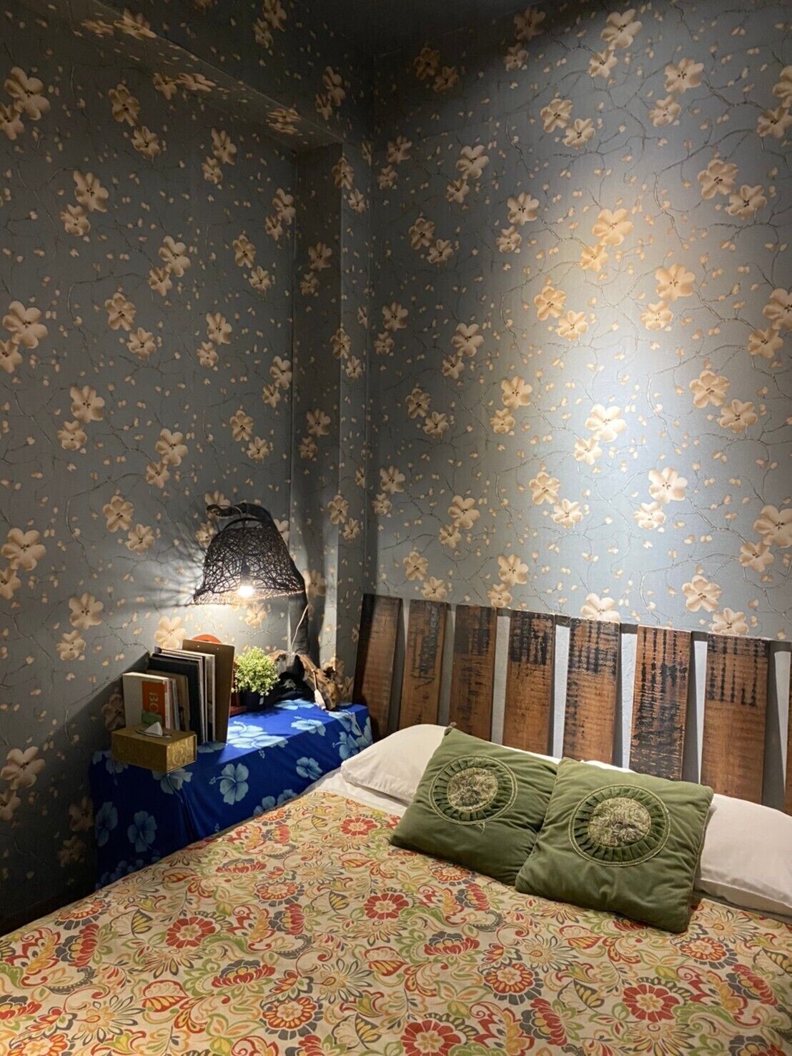 A bedroom with floral wallpaper, a bed with patterned bedding, a nightstand with books and a lamp, and a wooden headboard.