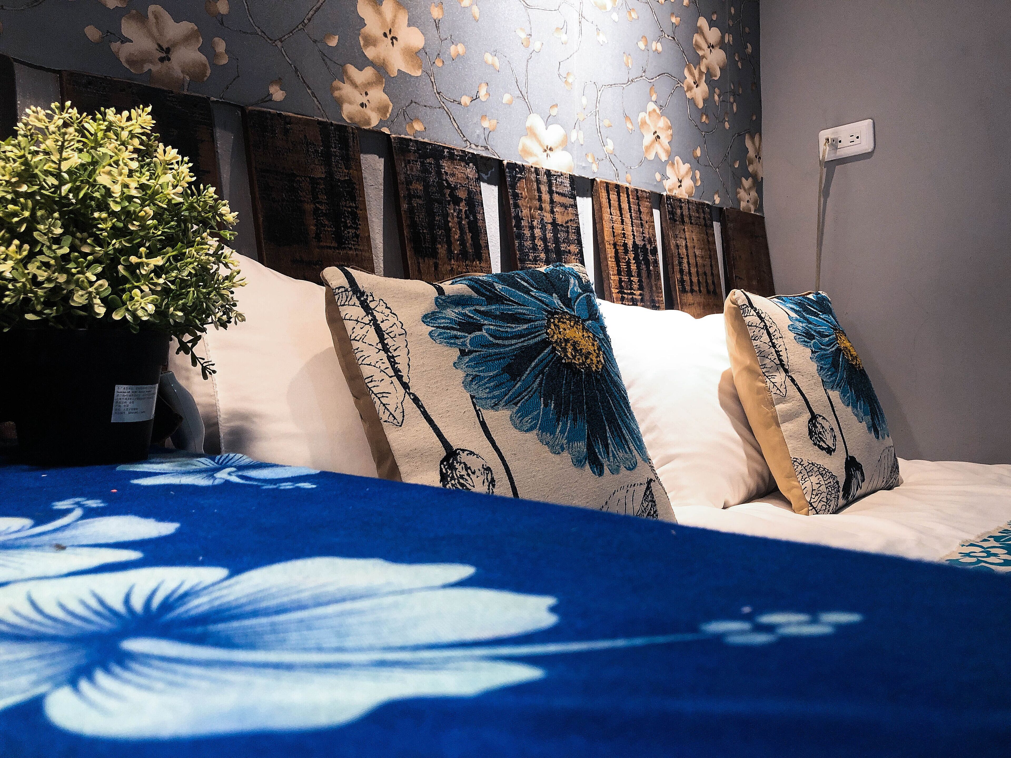 Una cama con almohadas estampadas con motivos florales, una planta en maceta y una pared con diseño de papel tapiz floral.