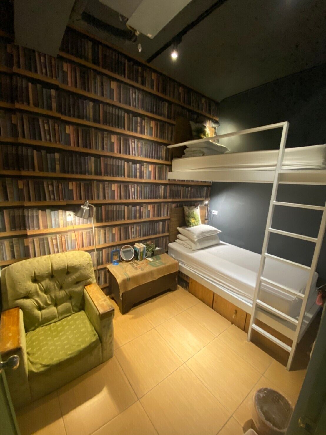 Een knus, klein kamertje met een stapelbed, een groene fauteuil, een boekenplank vol boeken en een tafeltje met een lamp.