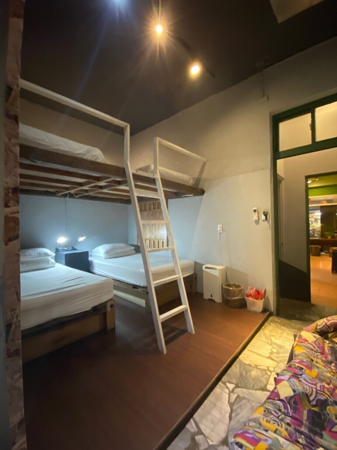 A dormitory room with bunk beds, a ladder, a bedside table, and a colorful bedspread.
