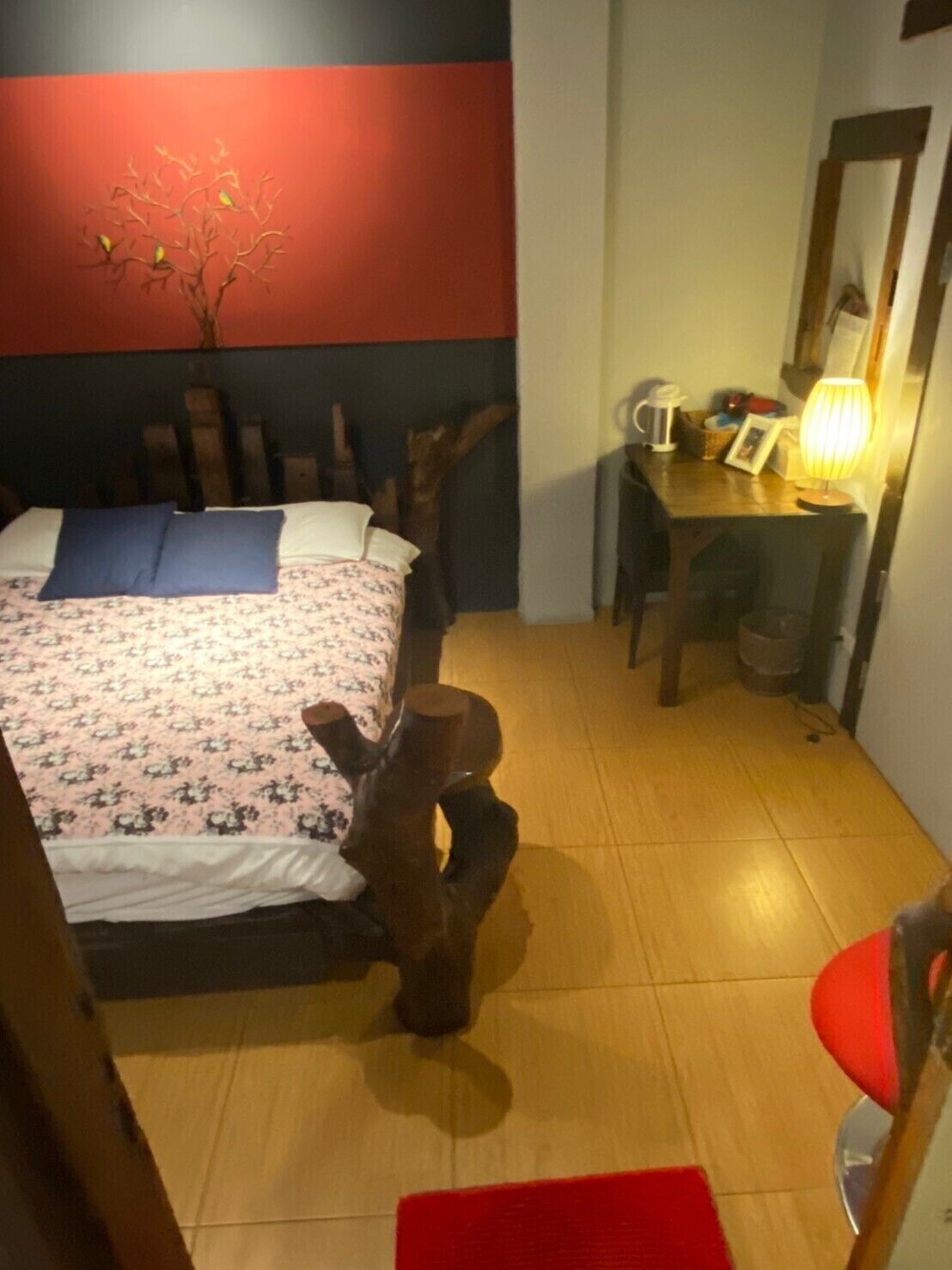 A bedroom with a bed, a wooden chair, a desk with a lamp, and a red wall.