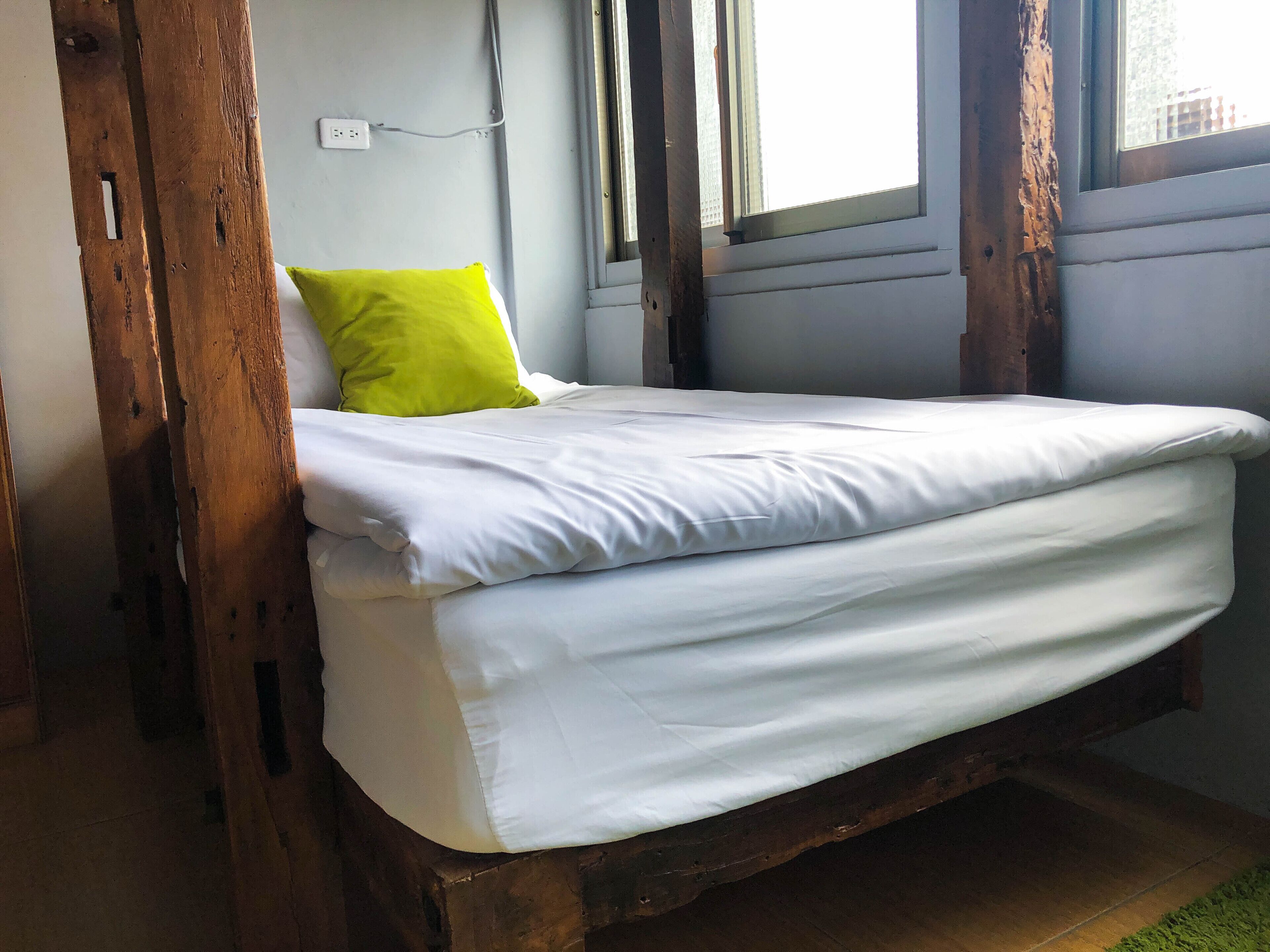 Un lit simple avec une literie blanche et un oreiller vert, dans une pièce dotée de poutres en bois et d’une fenêtre.