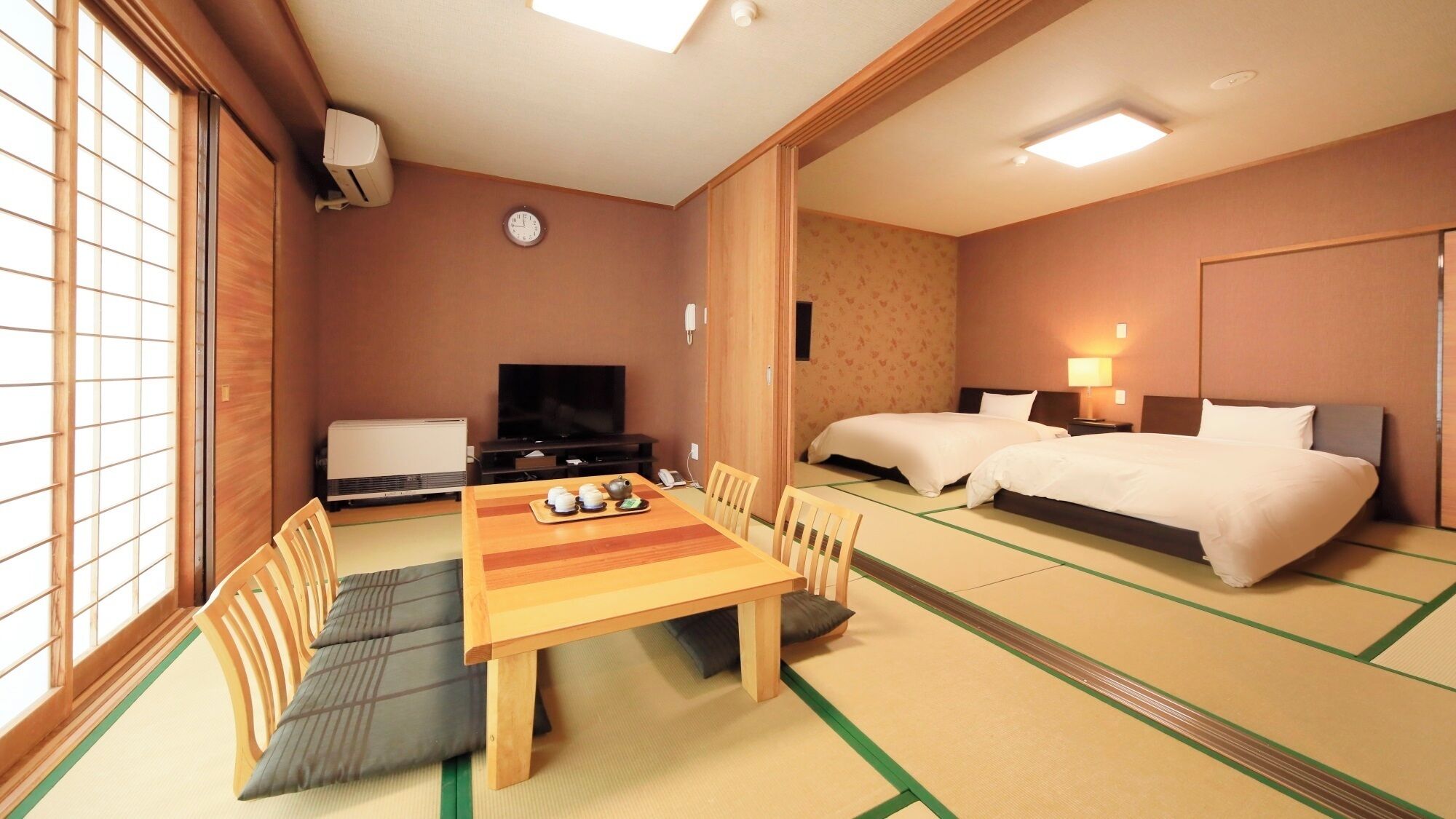 A hotel room with tatami flooring, a dining table with tea set, two beds, a television, and a clock on the wall.