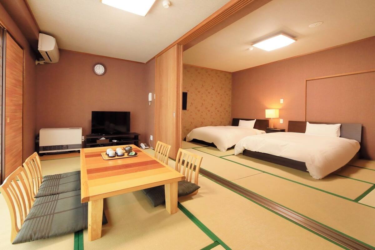 A hotel room with tatami flooring, a dining table with tea set, two beds, a television, and a clock on the wall.
